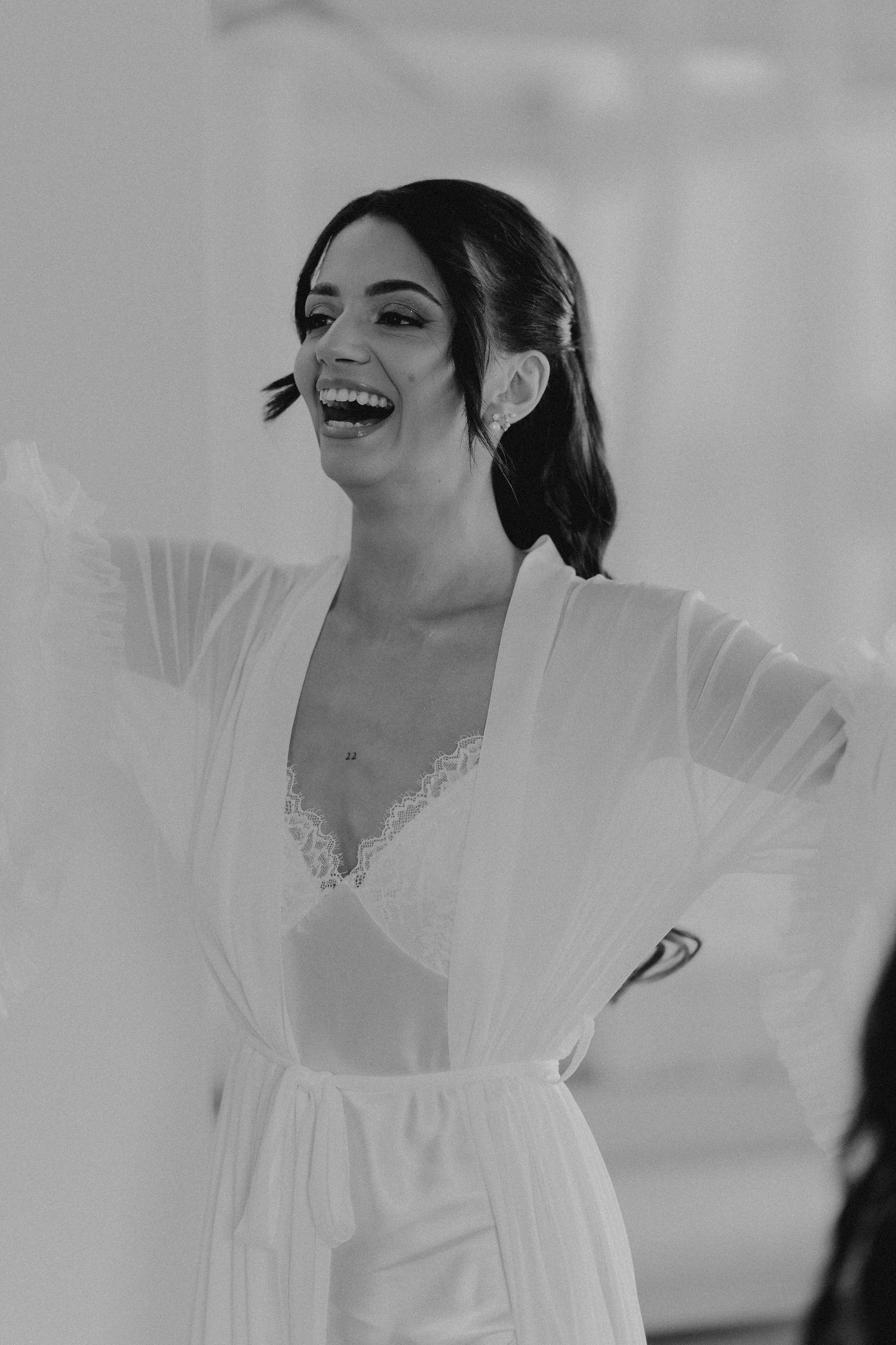 Black and white photo of a laughing bride in a white lace robe during wedding preparations.