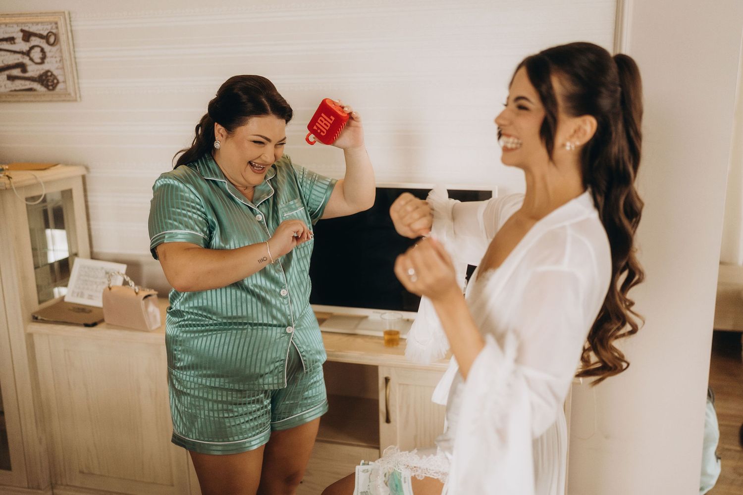 Two women in robes laugh and celebrate together in a bright room during bridal preparations.