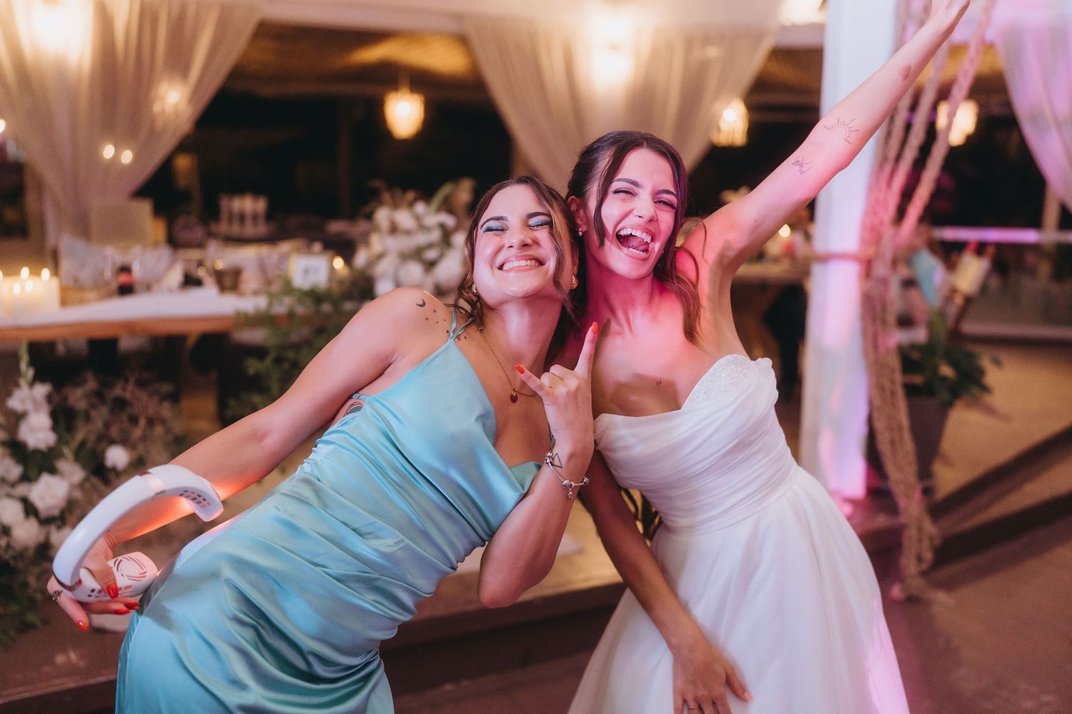 Bride in white strapless gown and bridesmaid in blue dress laughing and dancing together at wedding reception.