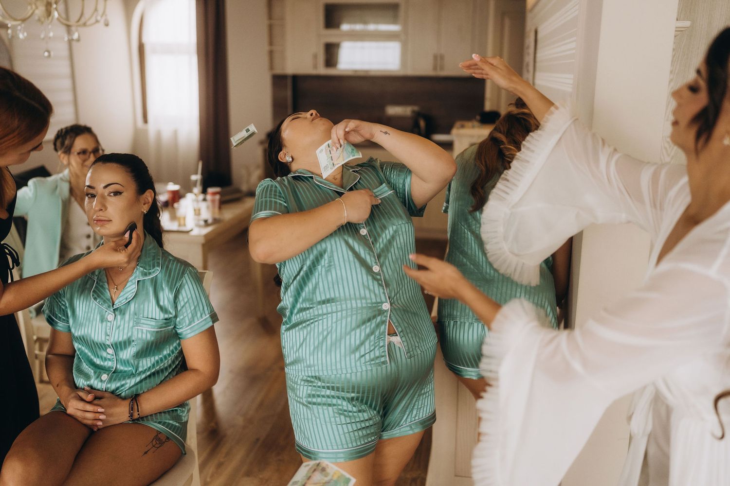 Bridesmaids in teal satin pajamas getting hair and makeup done together in a bright room before a wedding.