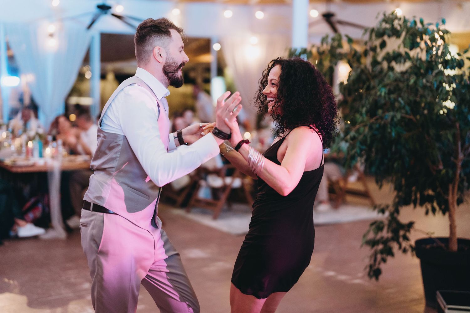 Couple dancing joyfully at a wedding reception, woman in black dress laughing with man in gray vest.