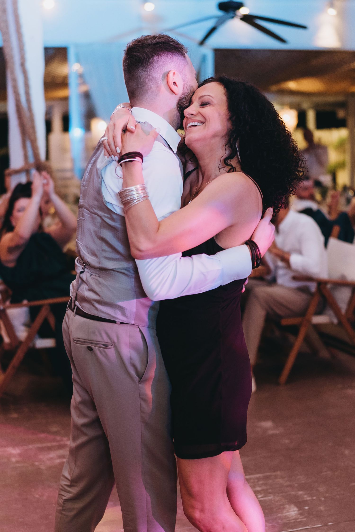 A groom and his mother share a heartfelt dance at a wedding reception, smiling and embracing on the dance floor.