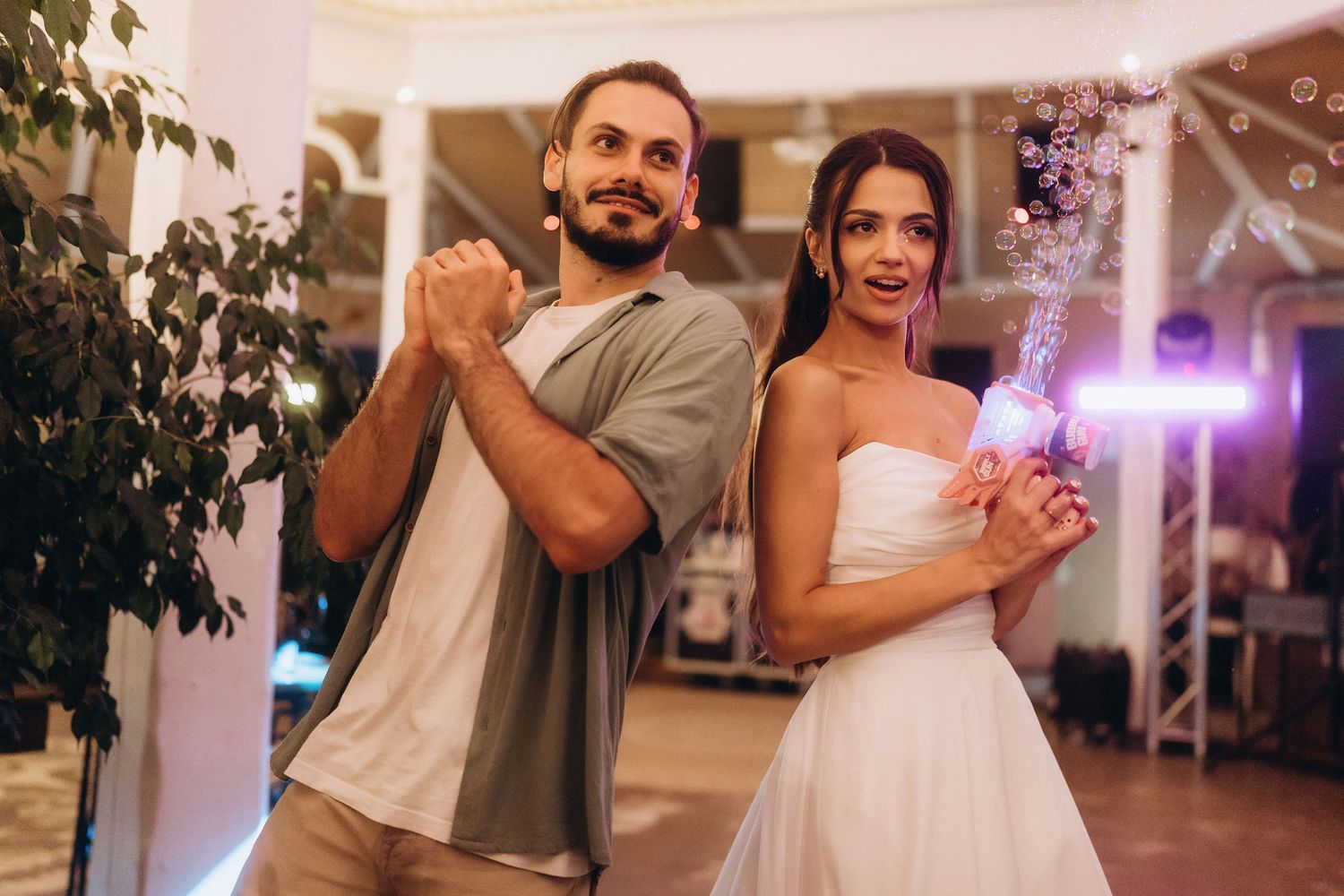 A smiling couple dancing at a wedding reception, bride in white dress holding flowers under purple lights.