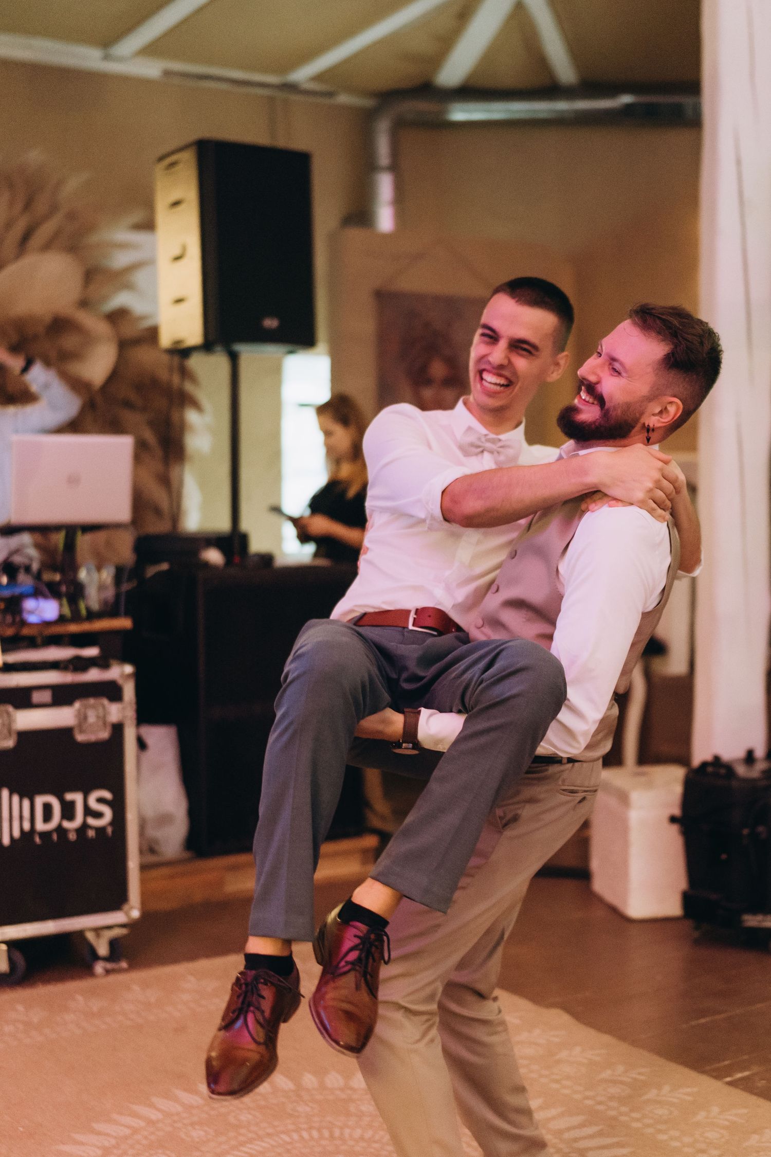 Two grooms share a joyful dance, one lifting the other, at their wedding reception.