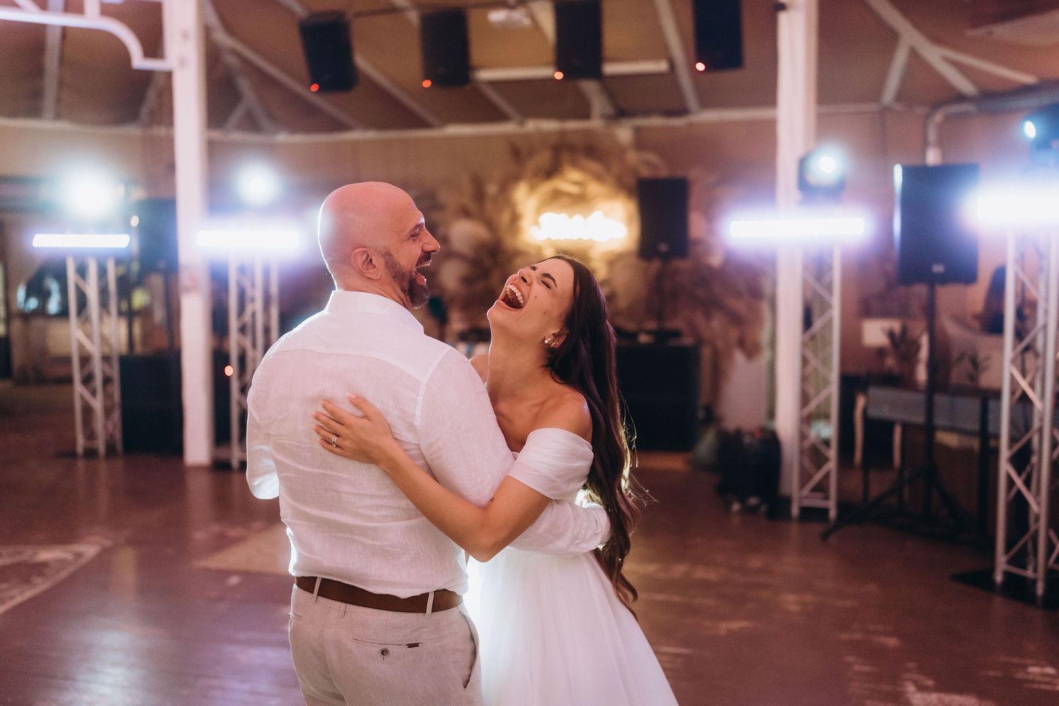 Bride and groom share a joyful first dance, laughing together under bright lights at their wedding reception.
