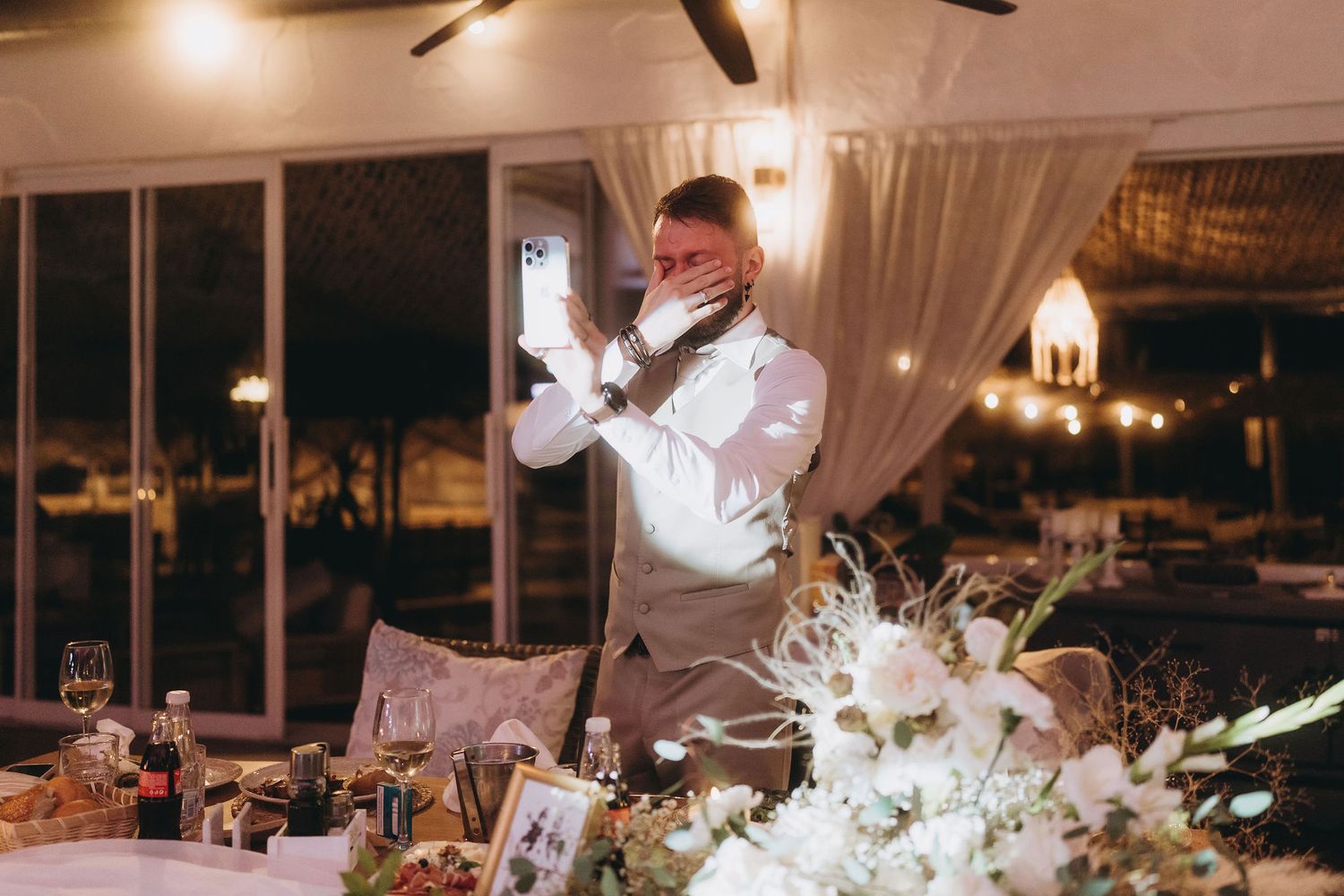 Groom adjusting his tie at elegant wedding reception with floral centerpieces and warm ambient lighting.