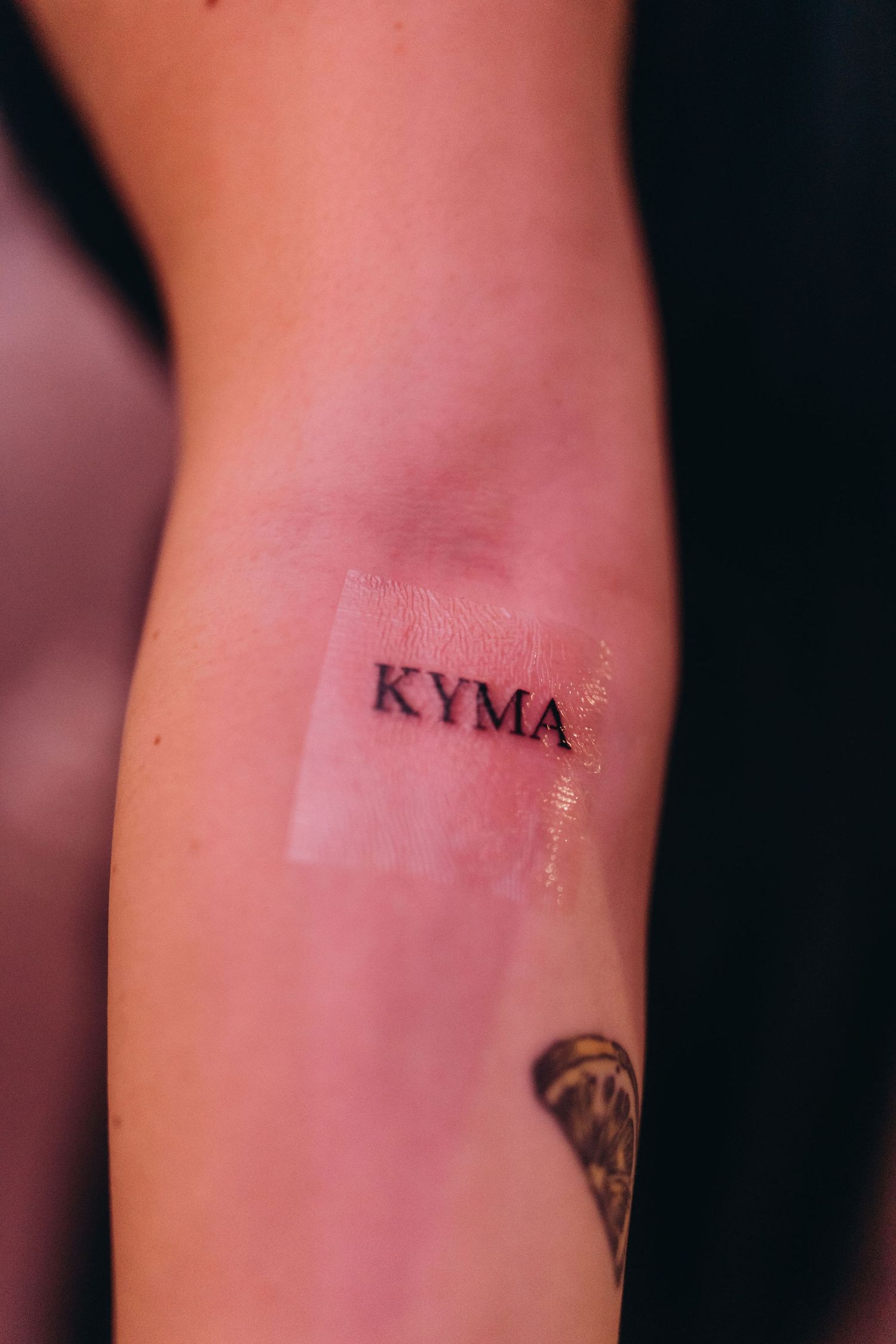 Close-up of a fresh "KYMA" minimalist text tattoo on a forearm, covered with protective film.