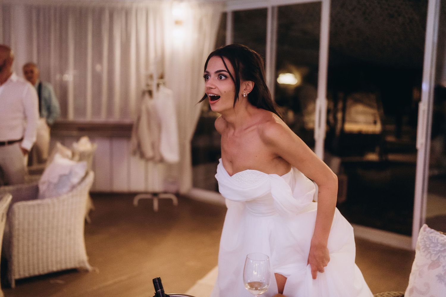 Surprised bride in white strapless wedding gown holding champagne glass at elegant indoor reception venue.