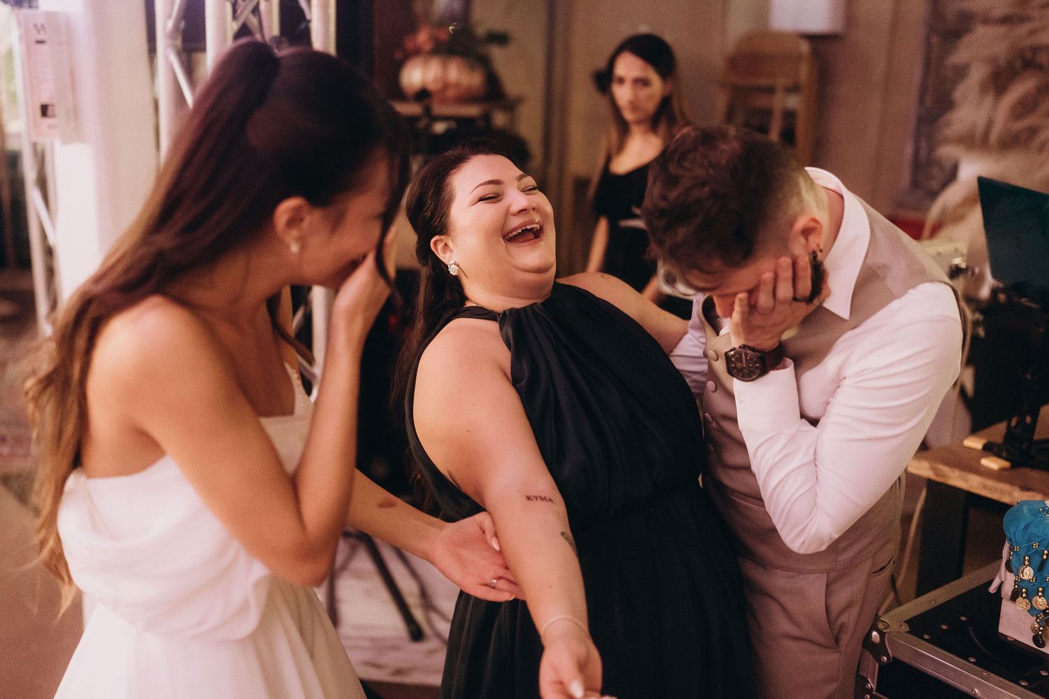 Three people laughing joyfully at a wedding, bride in white, woman in black dress, and groom covering his face.