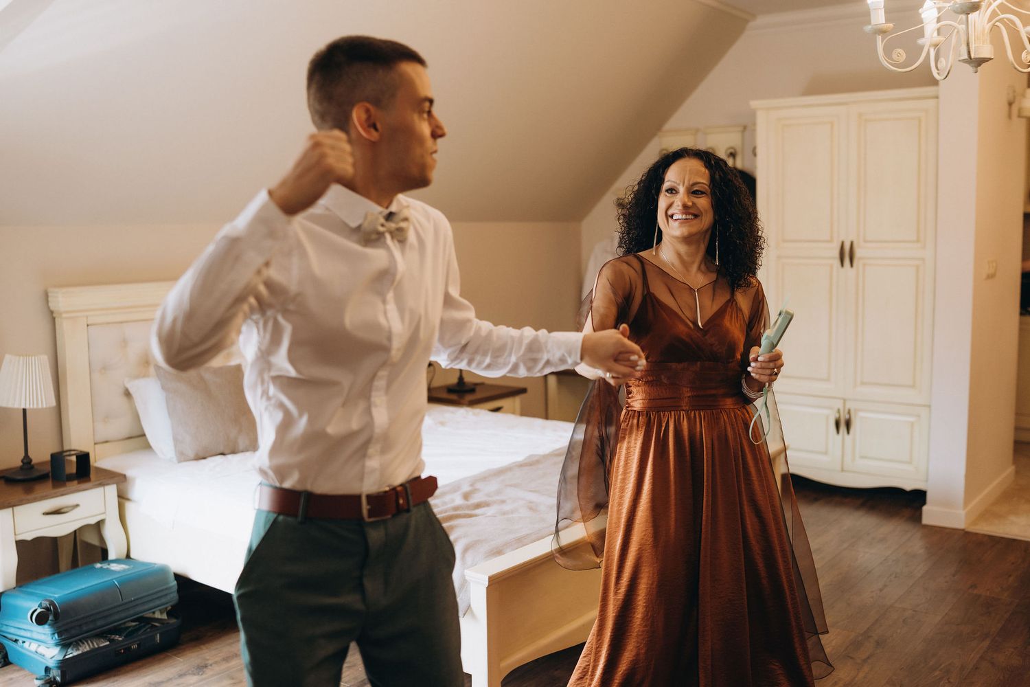 A couple getting ready for a formal event, woman in bronze gown helping man adjust his bow tie in elegant bedroom.