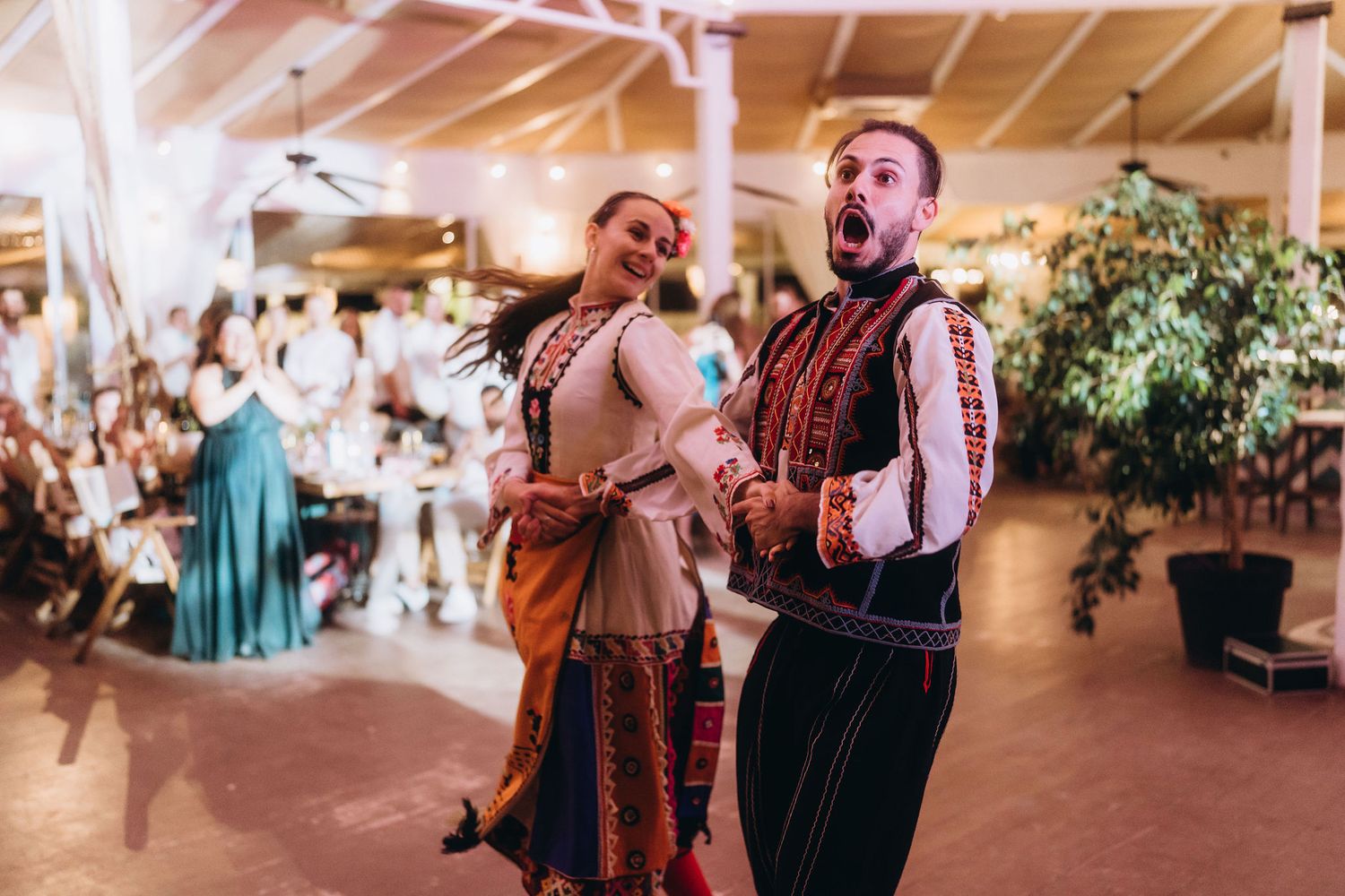 Two performers in traditional folk costumes dance energetically at a vibrant cultural celebration indoors.