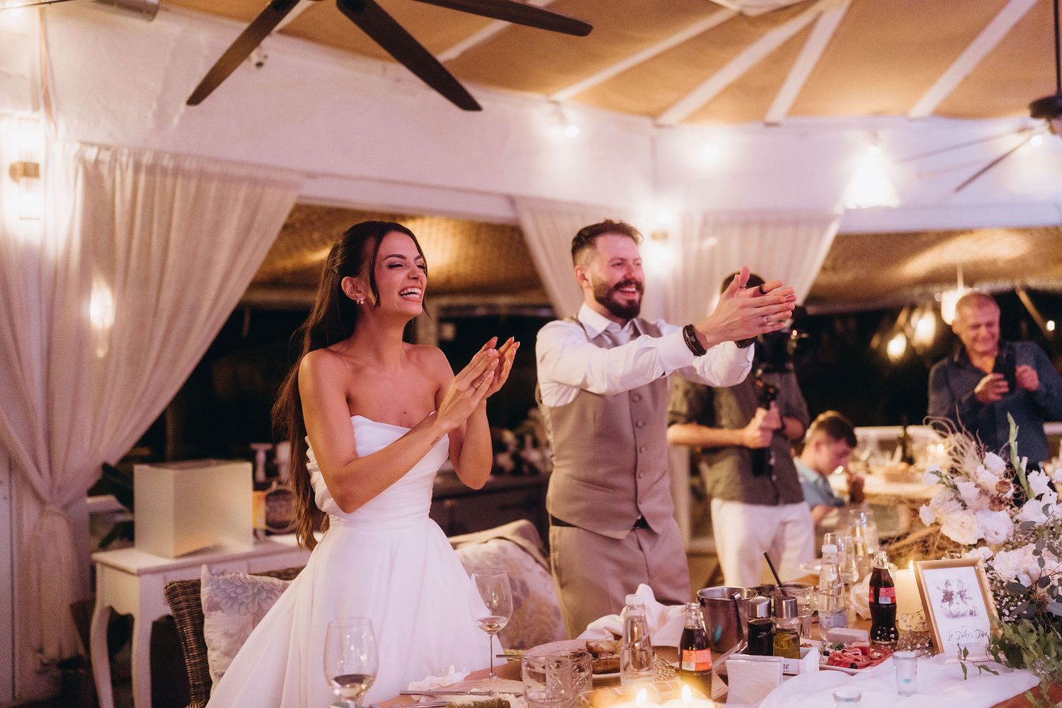 Bride and groom clapping and celebrating at their elegant wedding reception with warm string lights and floral decorations.