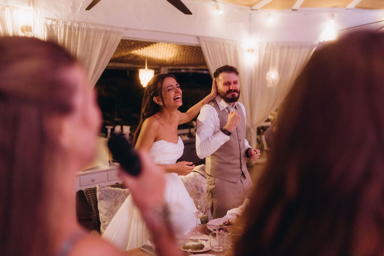 Bride and groom laughing joyfully during wedding reception with string lights and draped curtains in background.