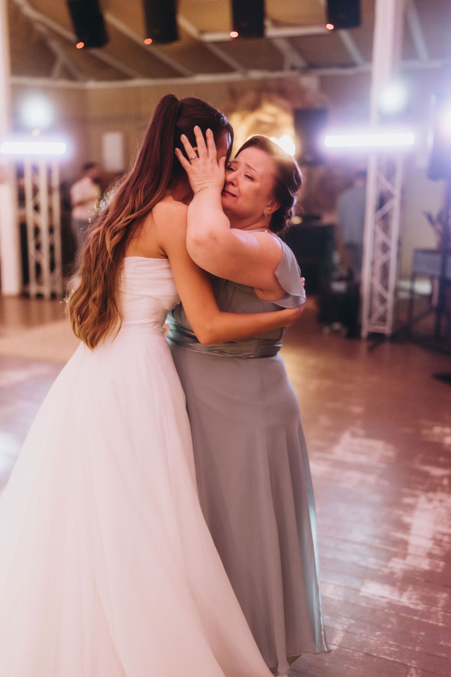 Bride and older woman share emotional dance floor embrace at wedding reception, woman tenderly cupping bride's face.