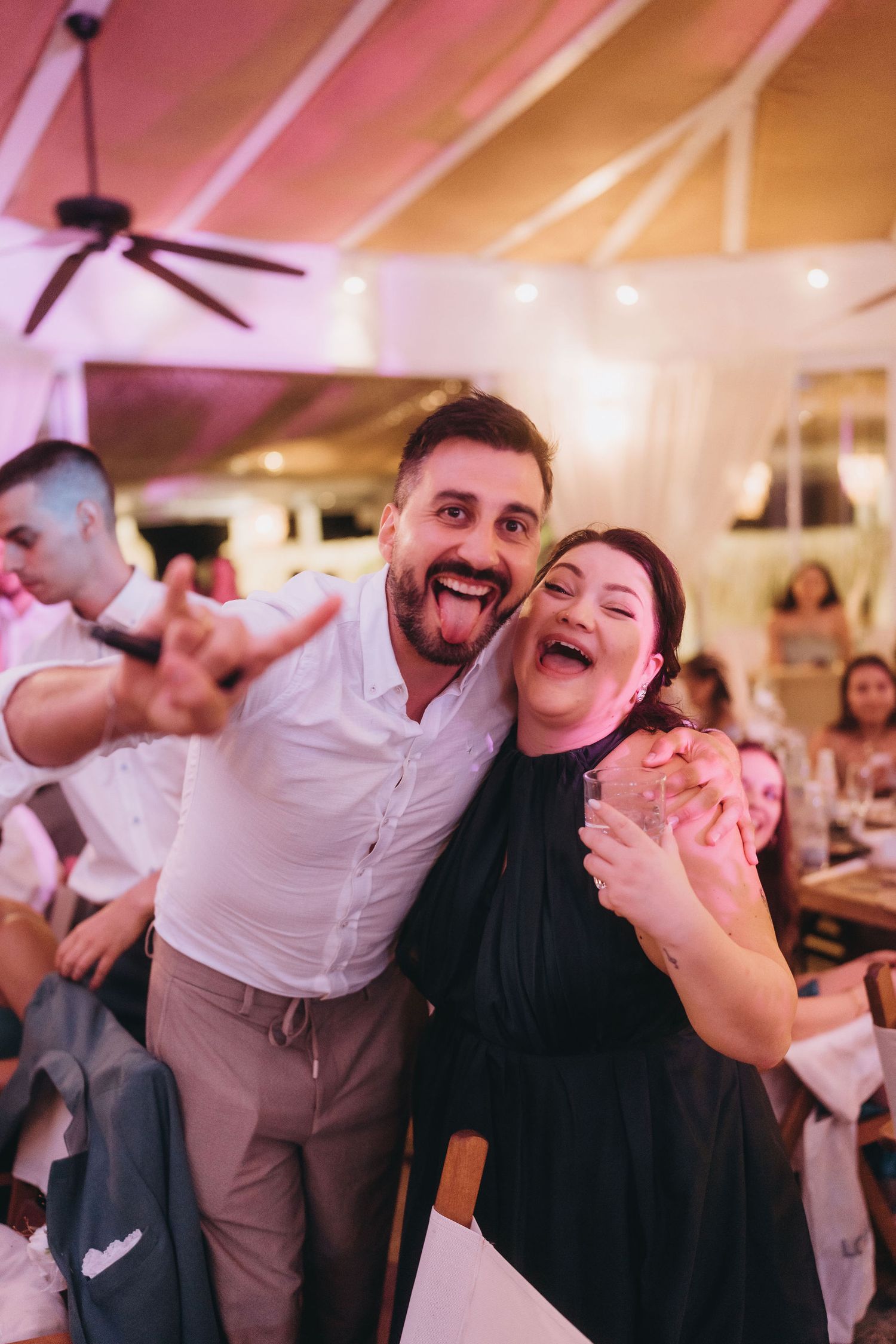 A joyful man and woman pose together at an indoor event, both smiling with tongues out in a lively venue.