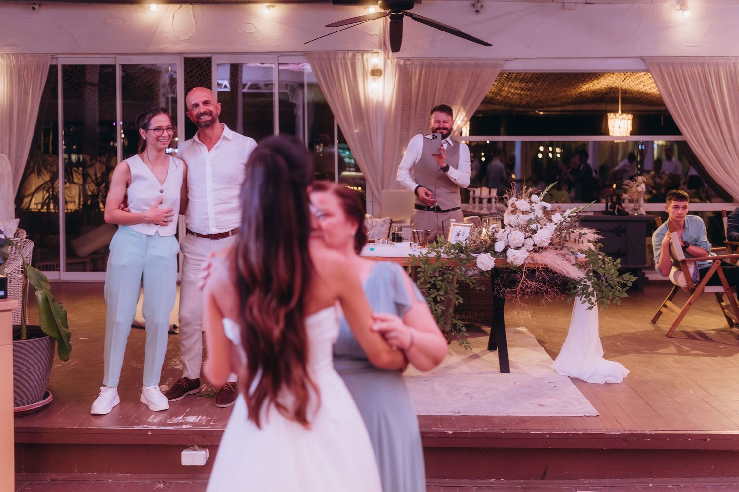 Guests watch a couple dance at an outdoor wedding reception with floral arrangements and string lights.