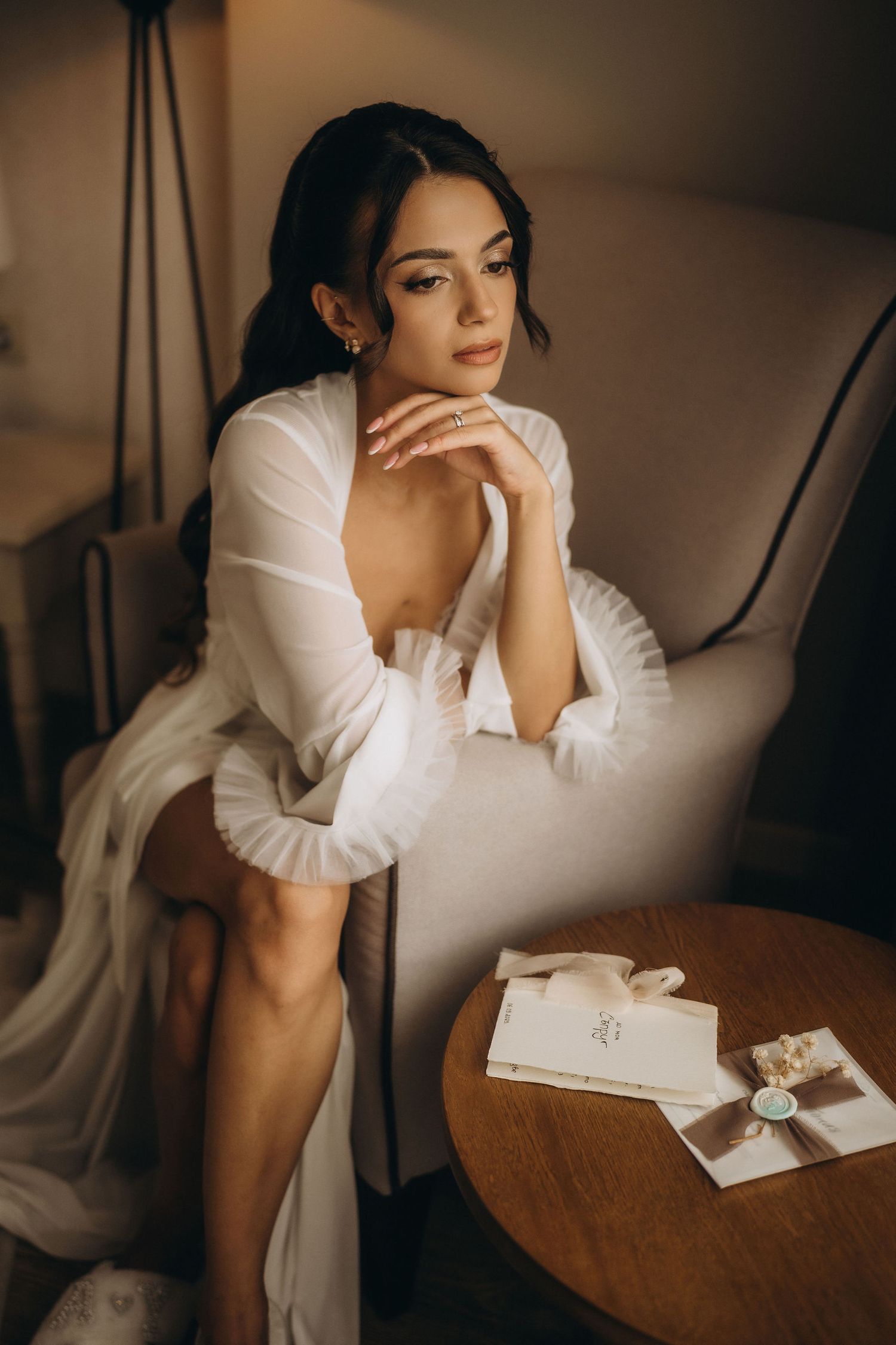 Elegant bride in white robe sitting in armchair, gazing thoughtfully, with wedding stationery on nearby table.