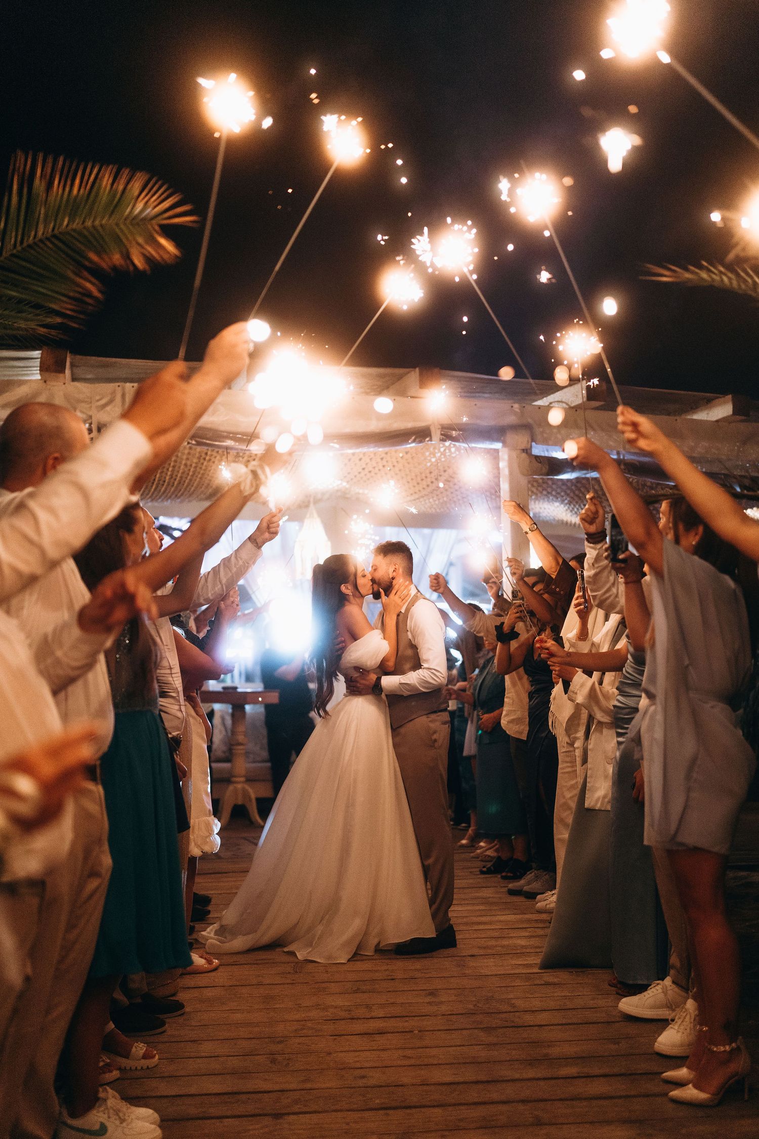 Bride and groom kissing under sparklers held by wedding guests during romantic outdoor night ceremony.