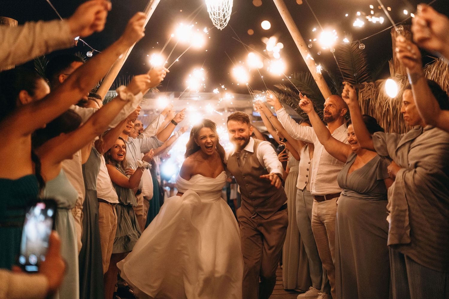 Bride and groom walk through sparkler tunnel surrounded by cheering wedding guests at night celebration.