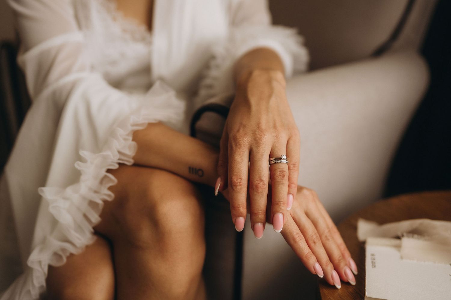 Bride in white robe sitting in chair showcasing elegant engagement ring with pink manicured nails.