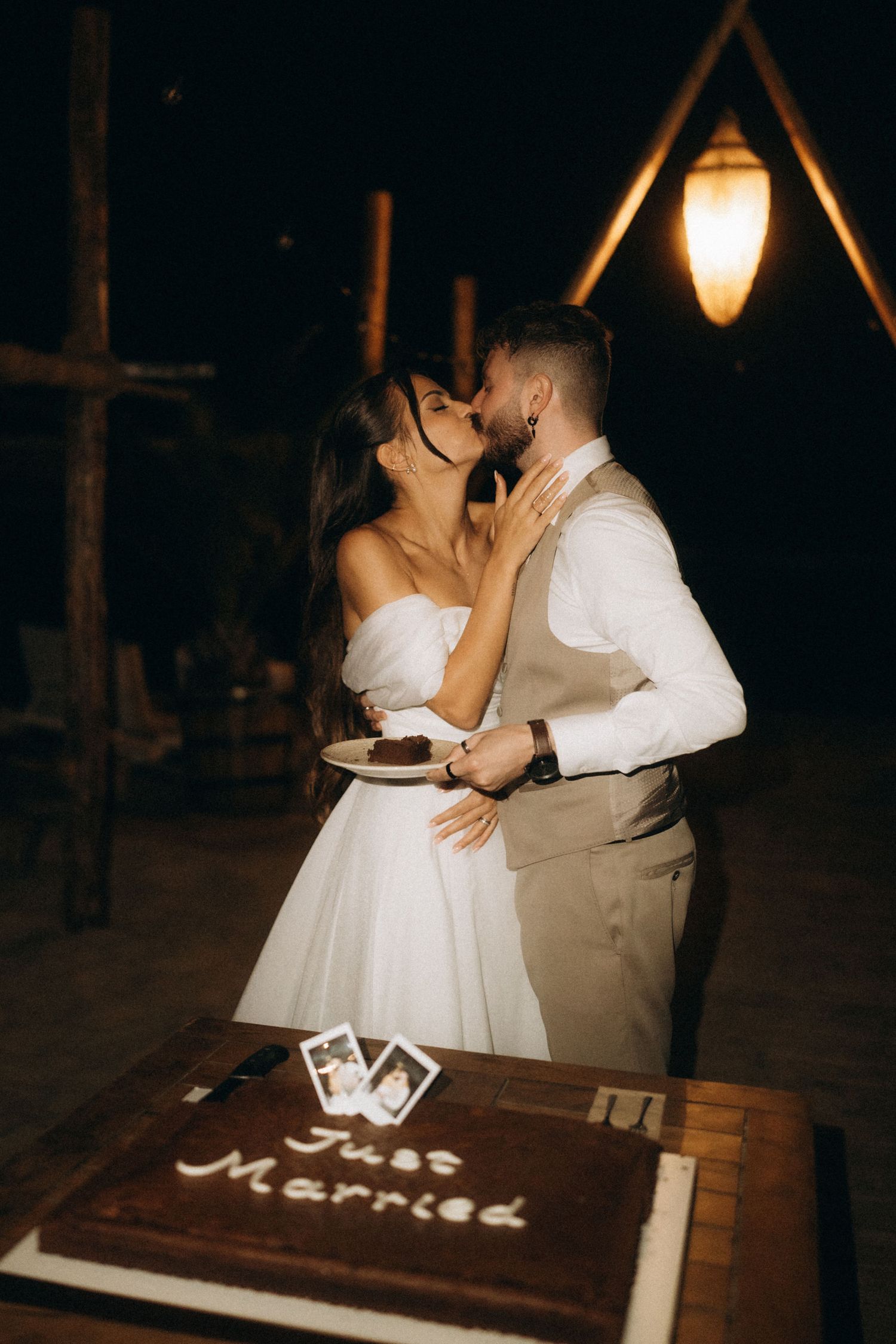 Newlyweds share a kiss beside their Just Married chocolate wedding cake at a romantic outdoor evening reception.