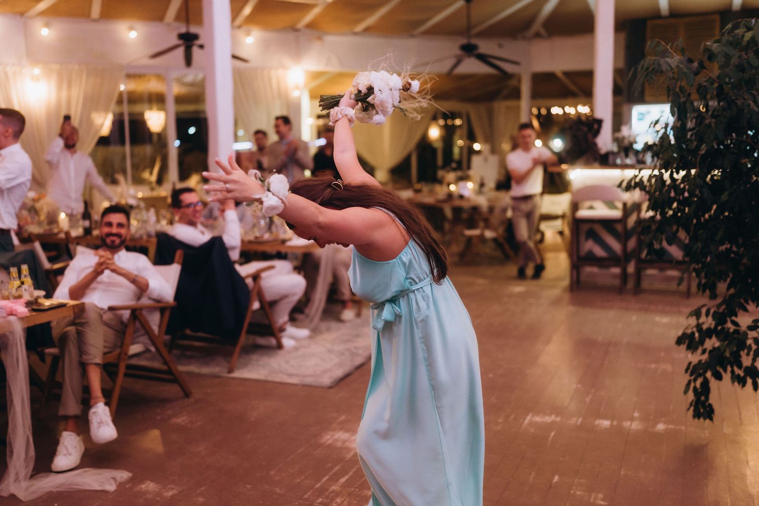 Bride in teal dress tossing wedding bouquet to excited guests at elegant indoor wedding reception.