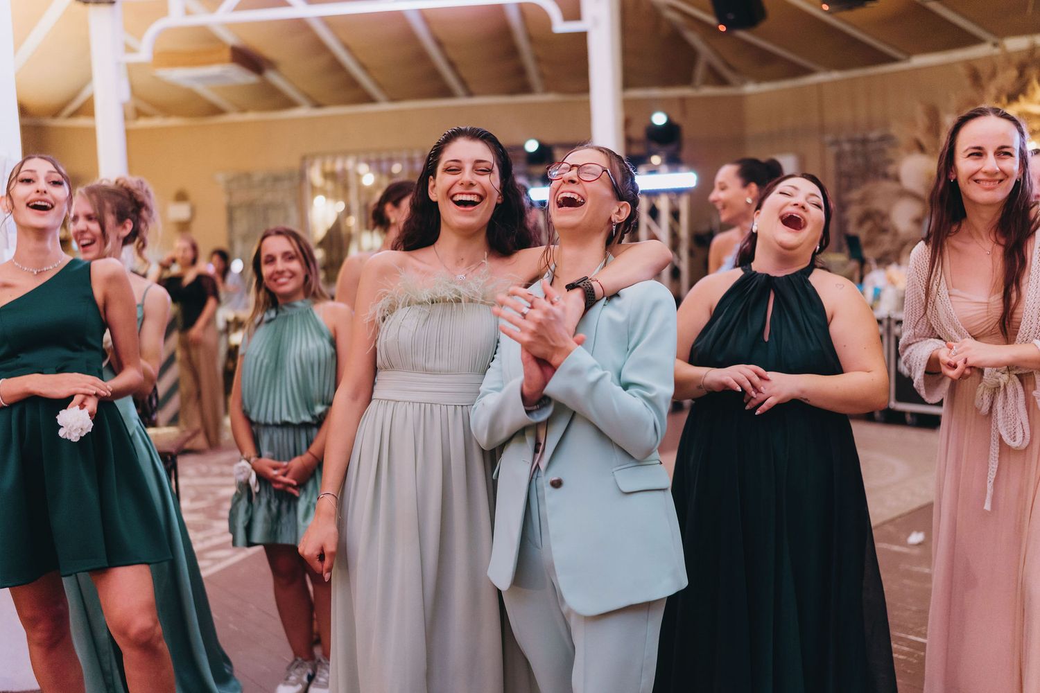 Group of women in elegant dresses laughing and dancing joyfully at a wedding reception celebration.