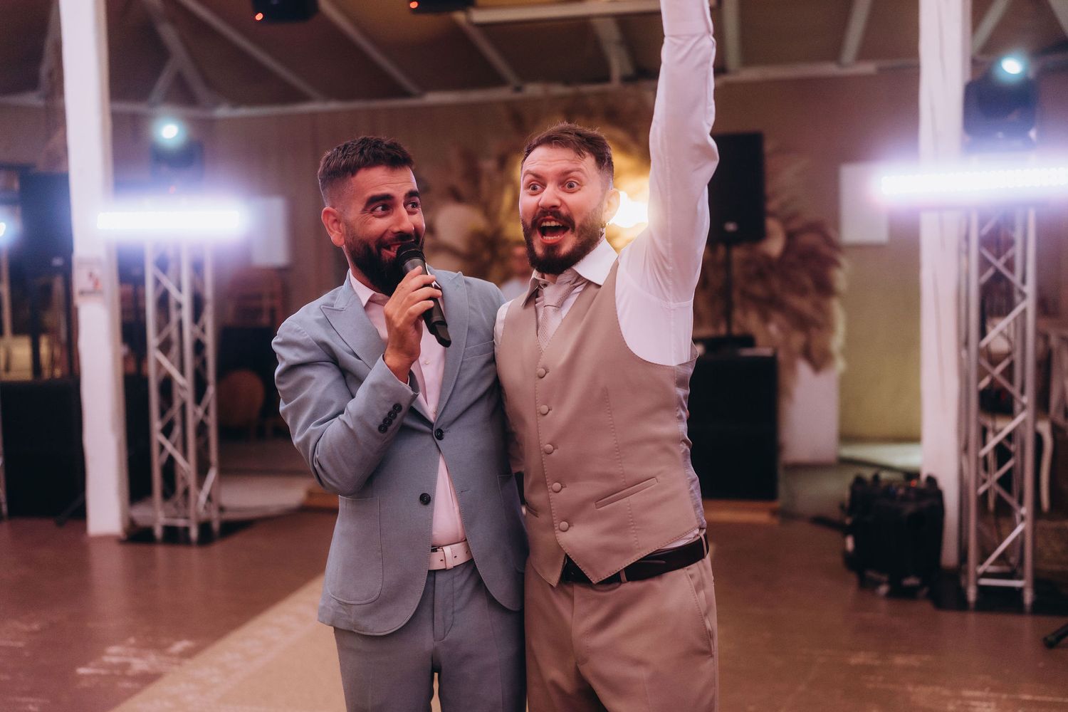 Two men in suits celebrate at a wedding reception, one holding a microphone and the other raising his hand.