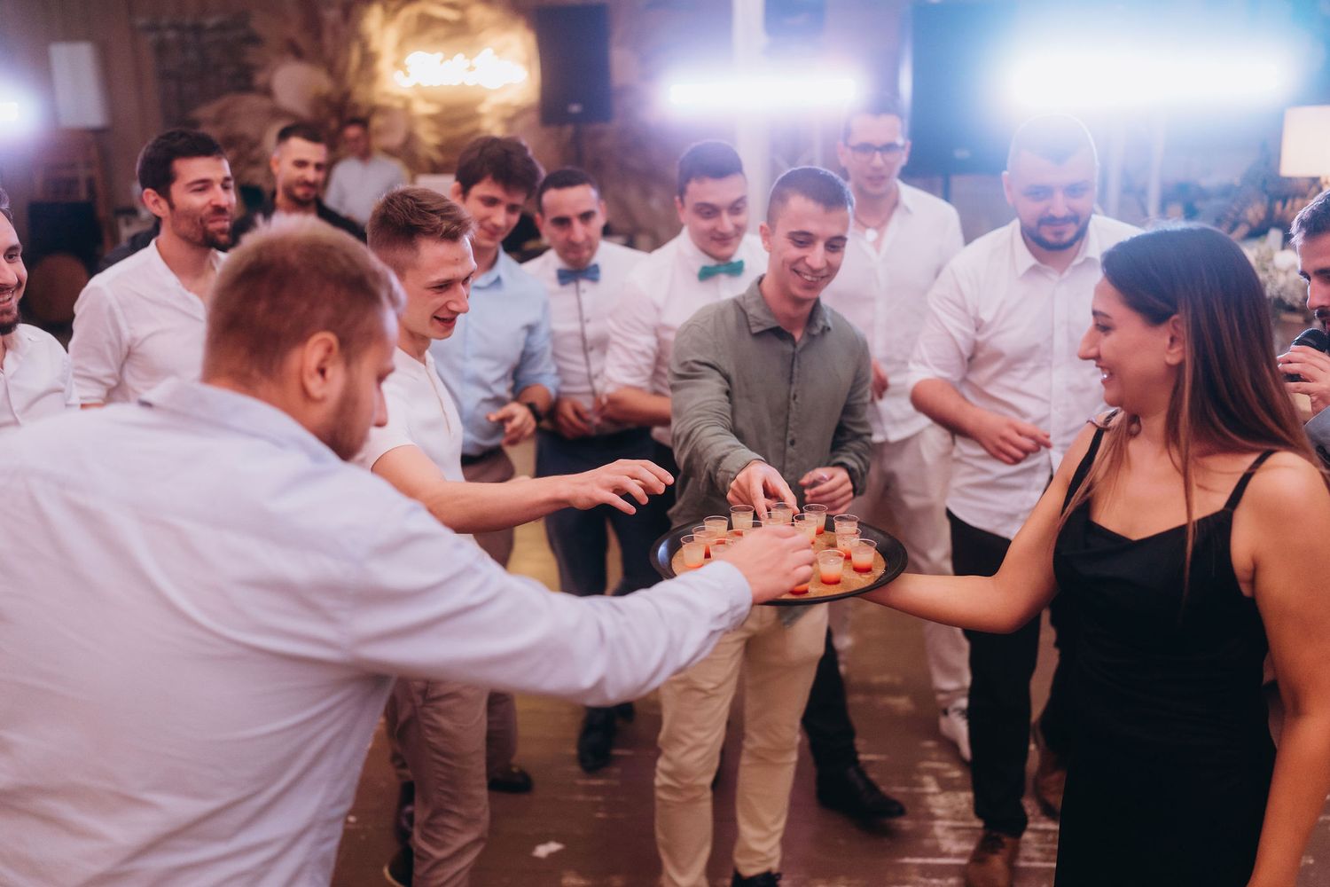 Wedding guests gather around a woman holding a tray of drinks at a lively reception party.