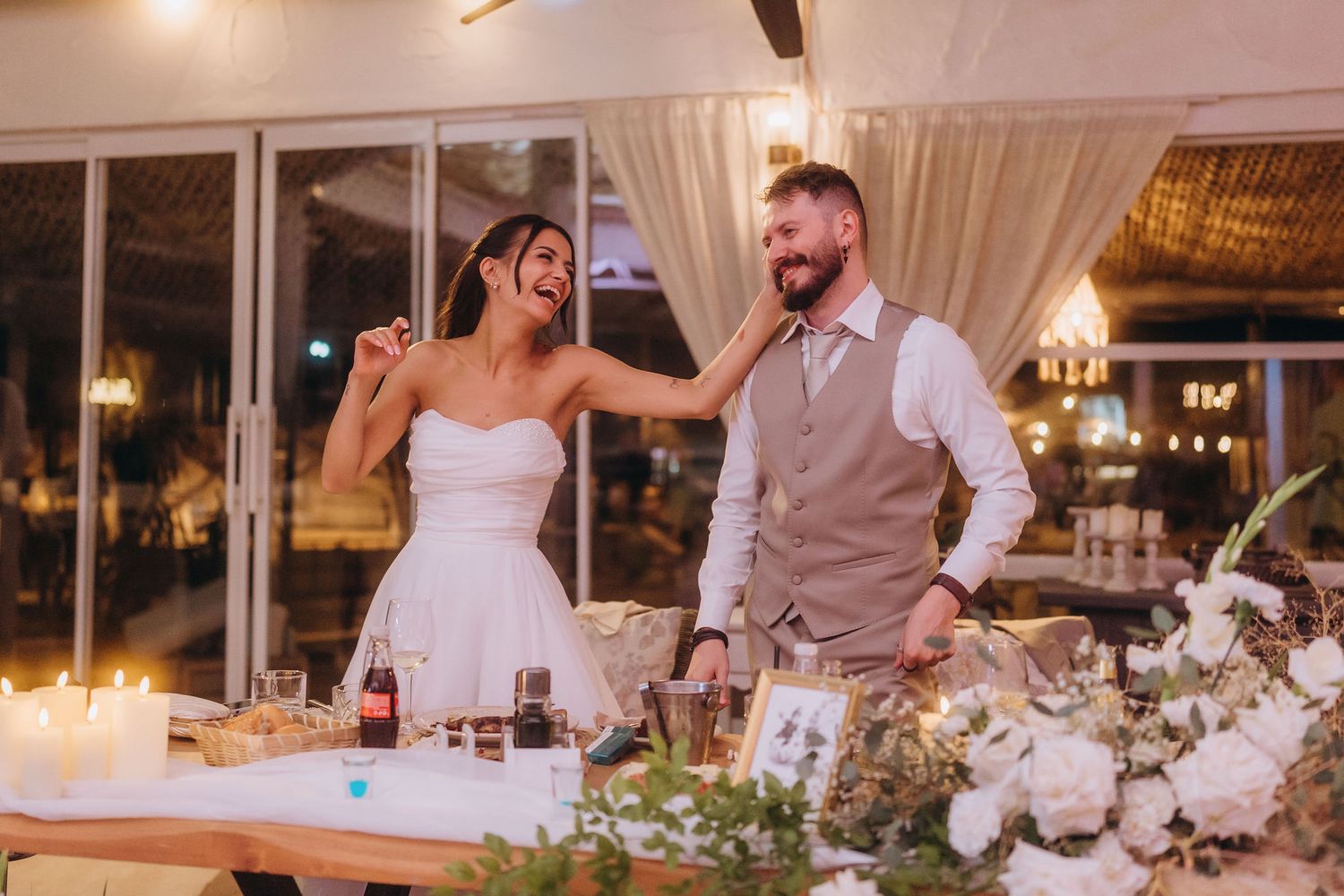A joyful bride and groom laugh together at their wedding reception table adorned with candles and white floral arrangements.