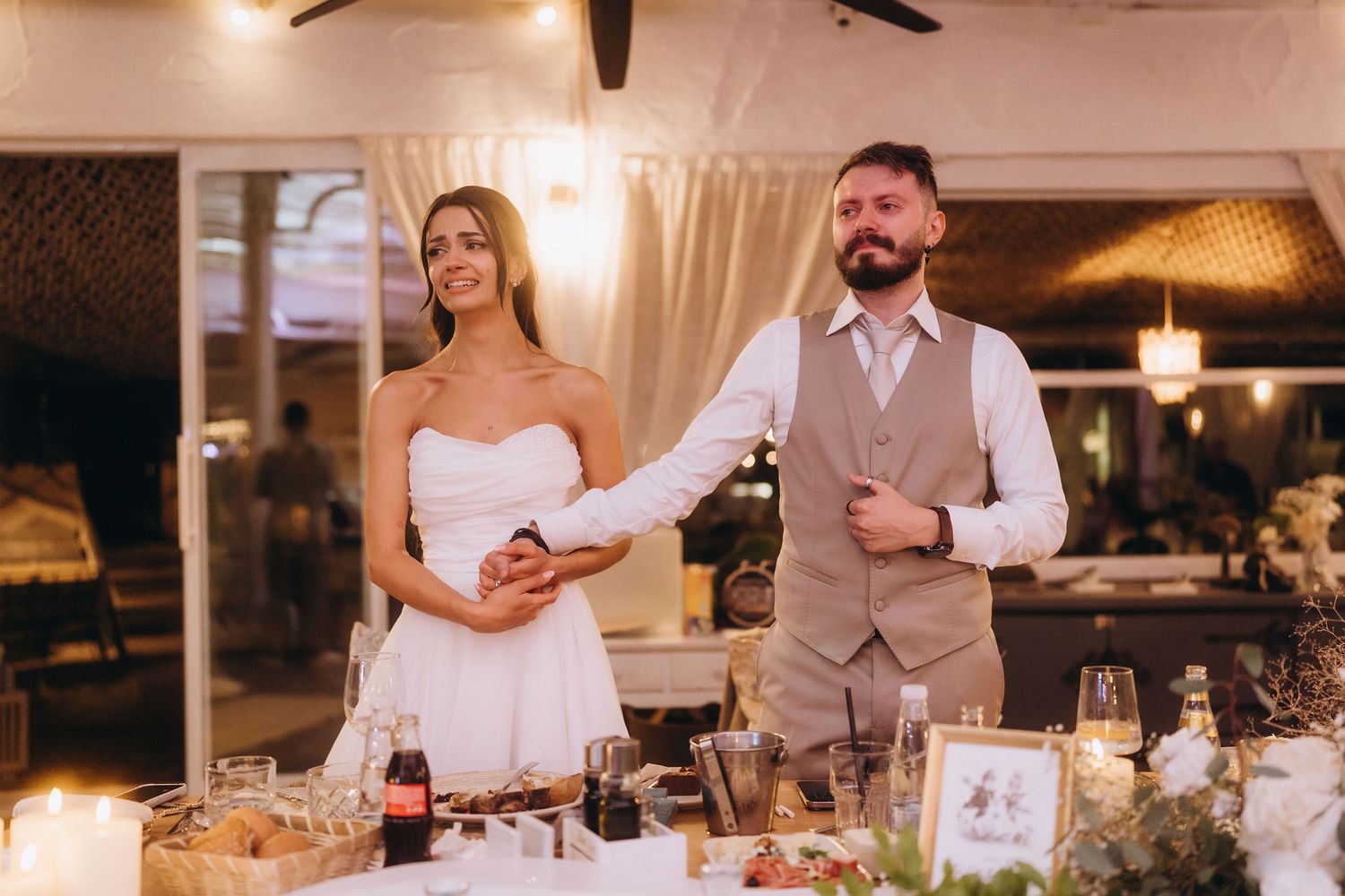 Bride and groom stand together at wedding reception table with candles and decorations in elegant venue.