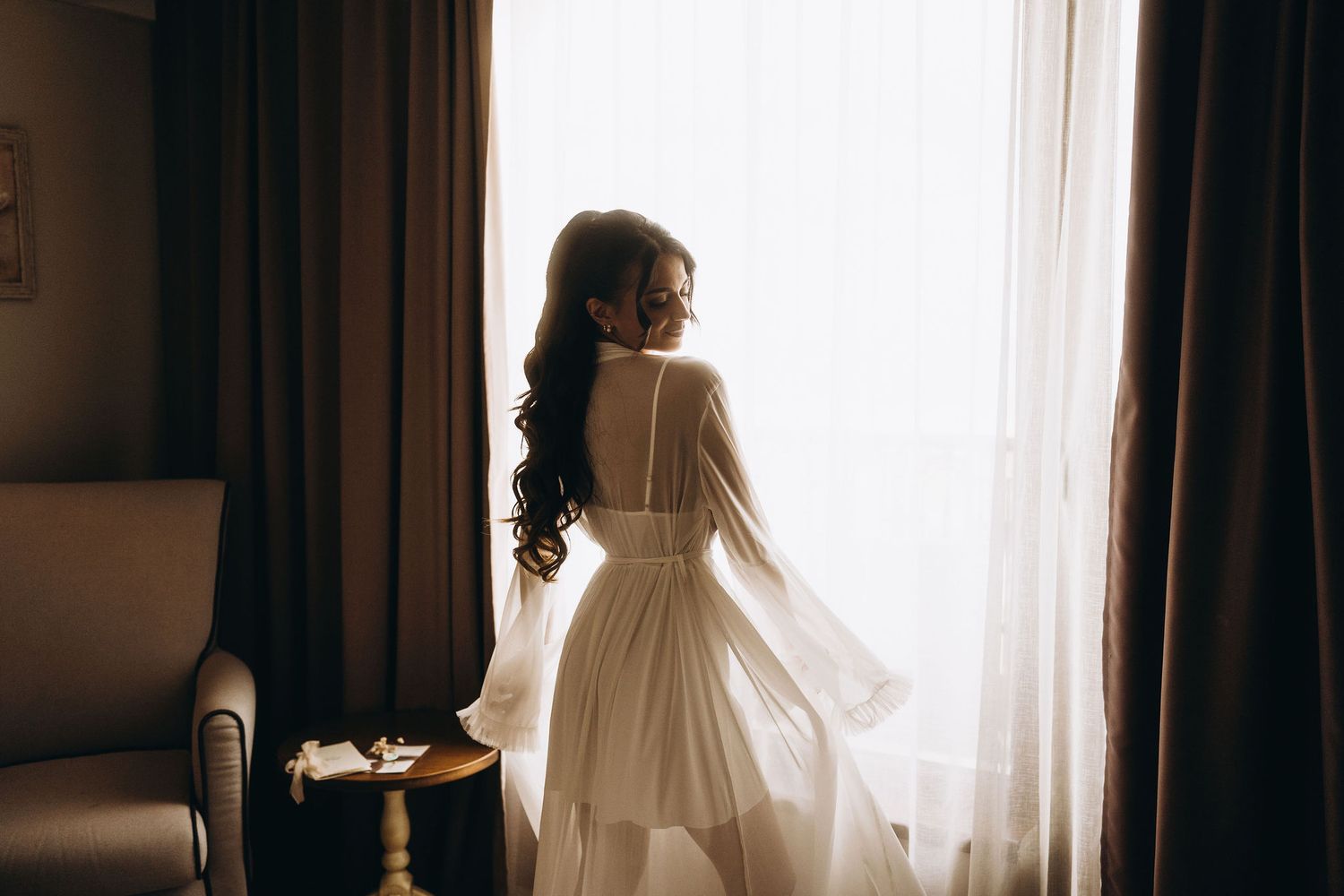 Elegant bride in a white flowing dress standing by sheer curtains in a softly lit room, viewed from behind.