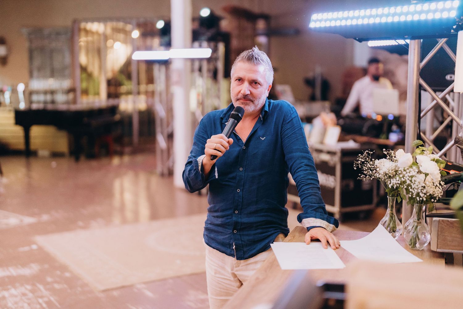 Middle-aged man with gray beard speaking into microphone at indoor event, wearing blue shirt near floral arrangement.