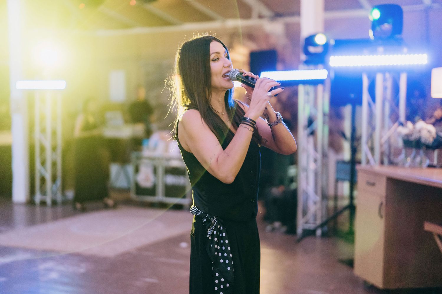 Woman in black dress singing into microphone on stage with bright lights at an indoor event.