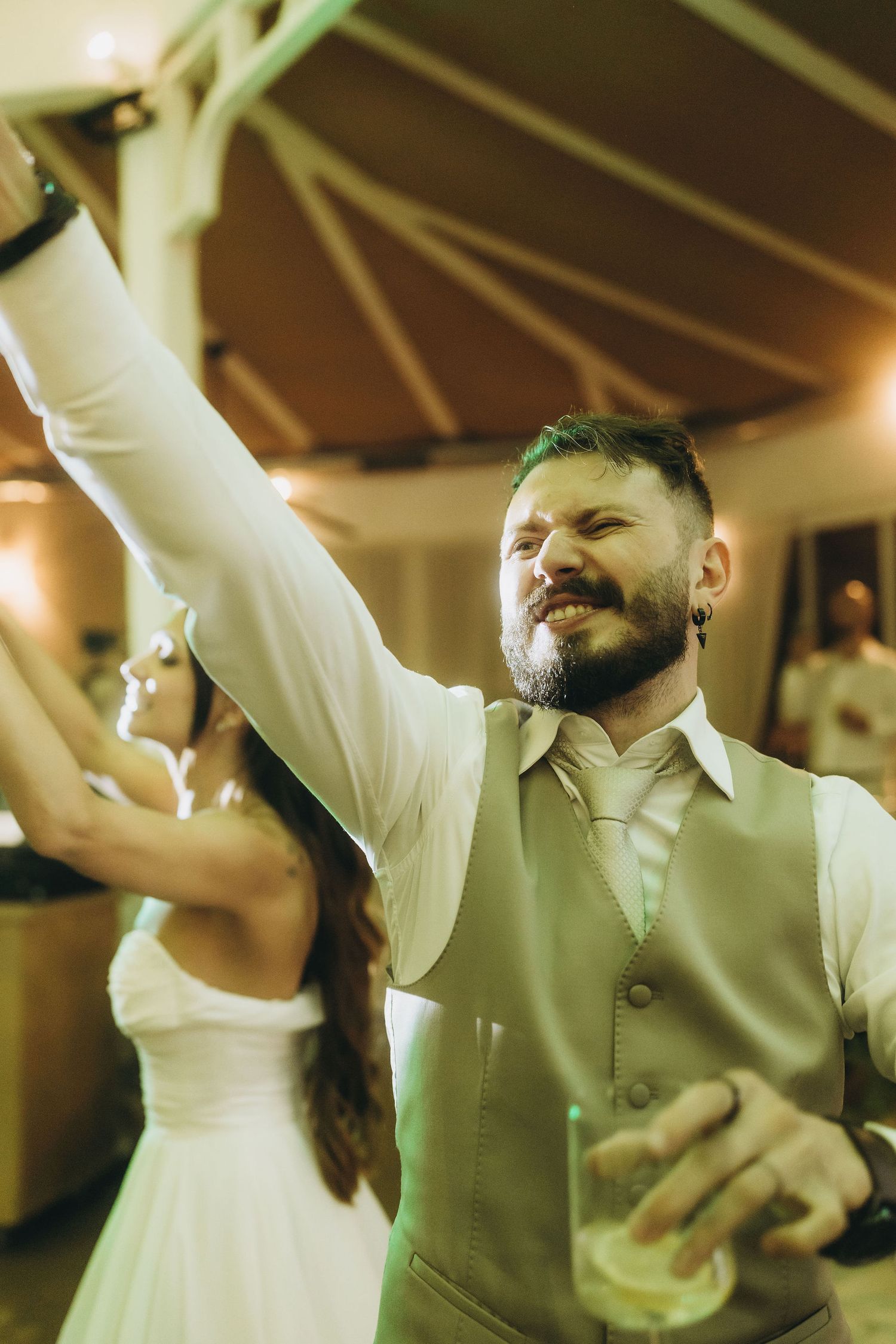 Bearded groom in white shirt and vest dancing joyfully at wedding reception, raising arm while holding drink.
