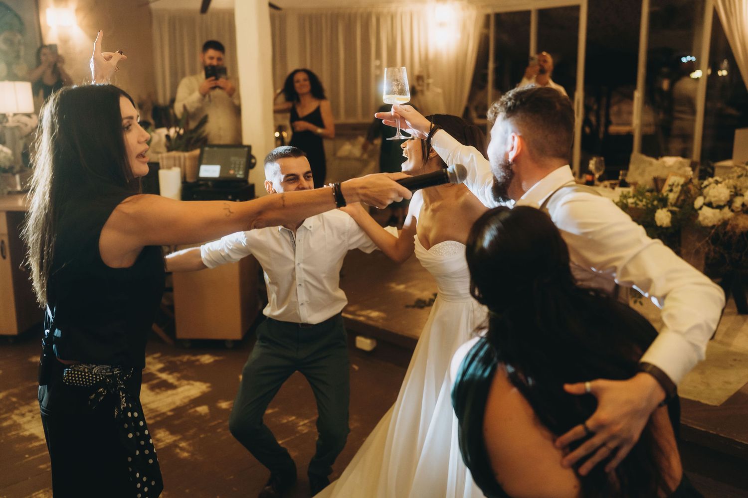 Wedding guests dancing and toasting with champagne glasses at a lively indoor wedding reception celebration.