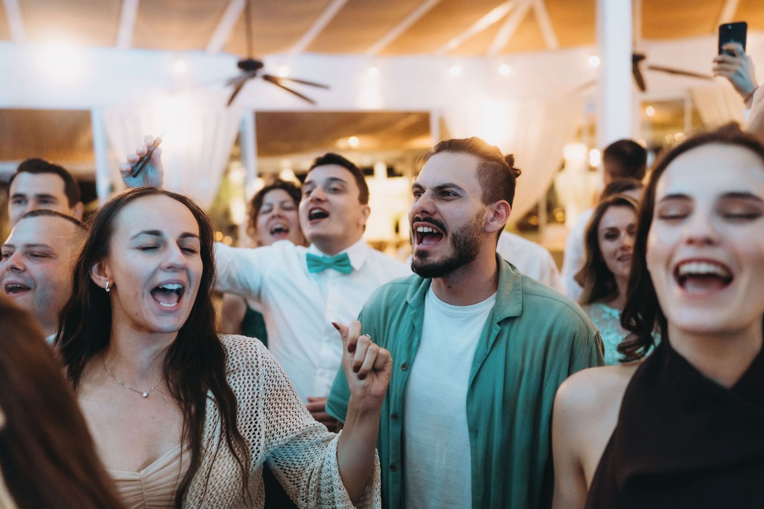 Guests laughing and singing joyfully at a lively wedding reception party indoors.