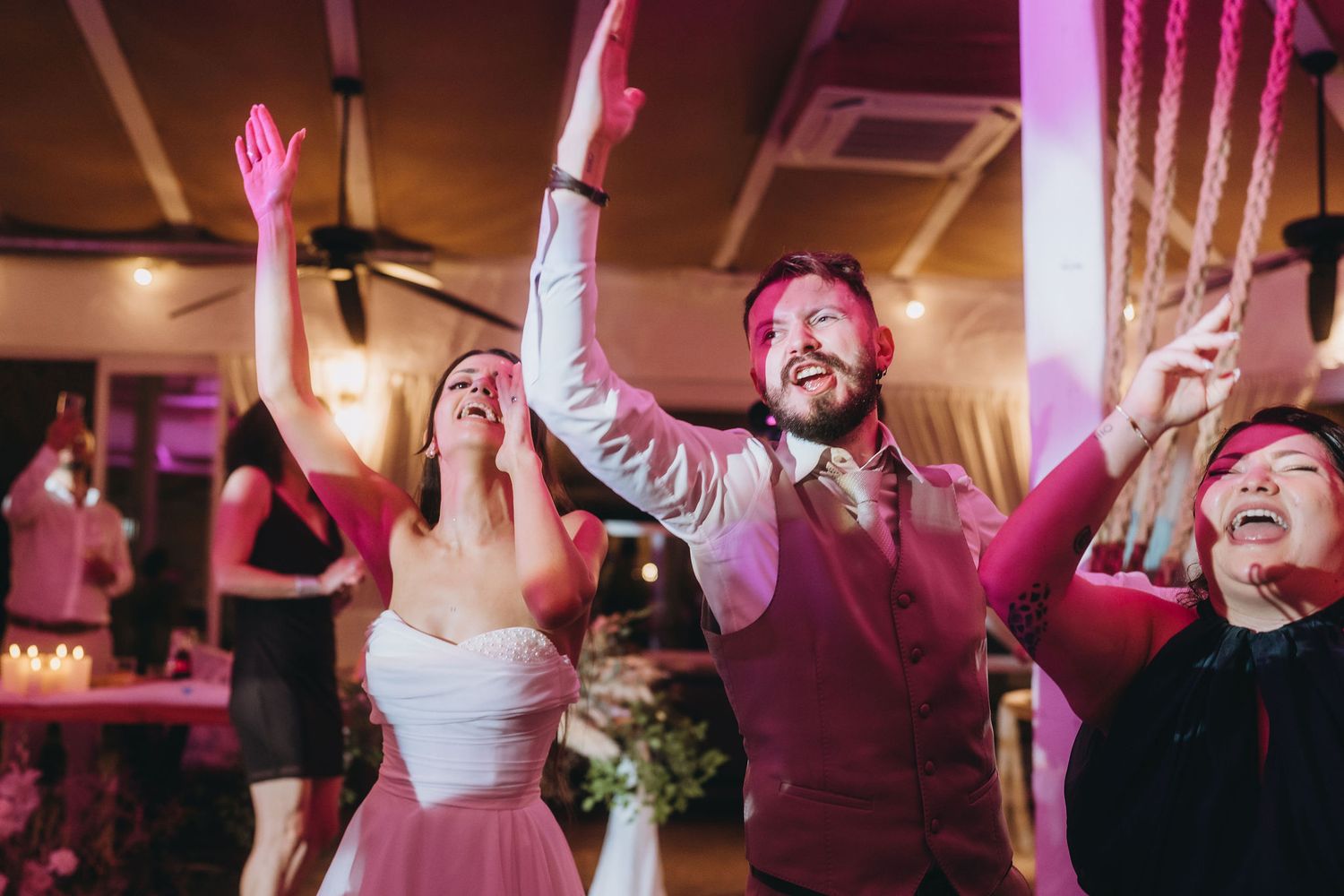 Joyful bride and groom dancing with guests at a romantic pink-lit wedding reception celebration.