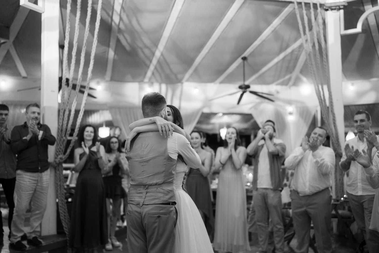Black and white photo of bride and groom sharing first dance while wedding guests clap and celebrate around them.