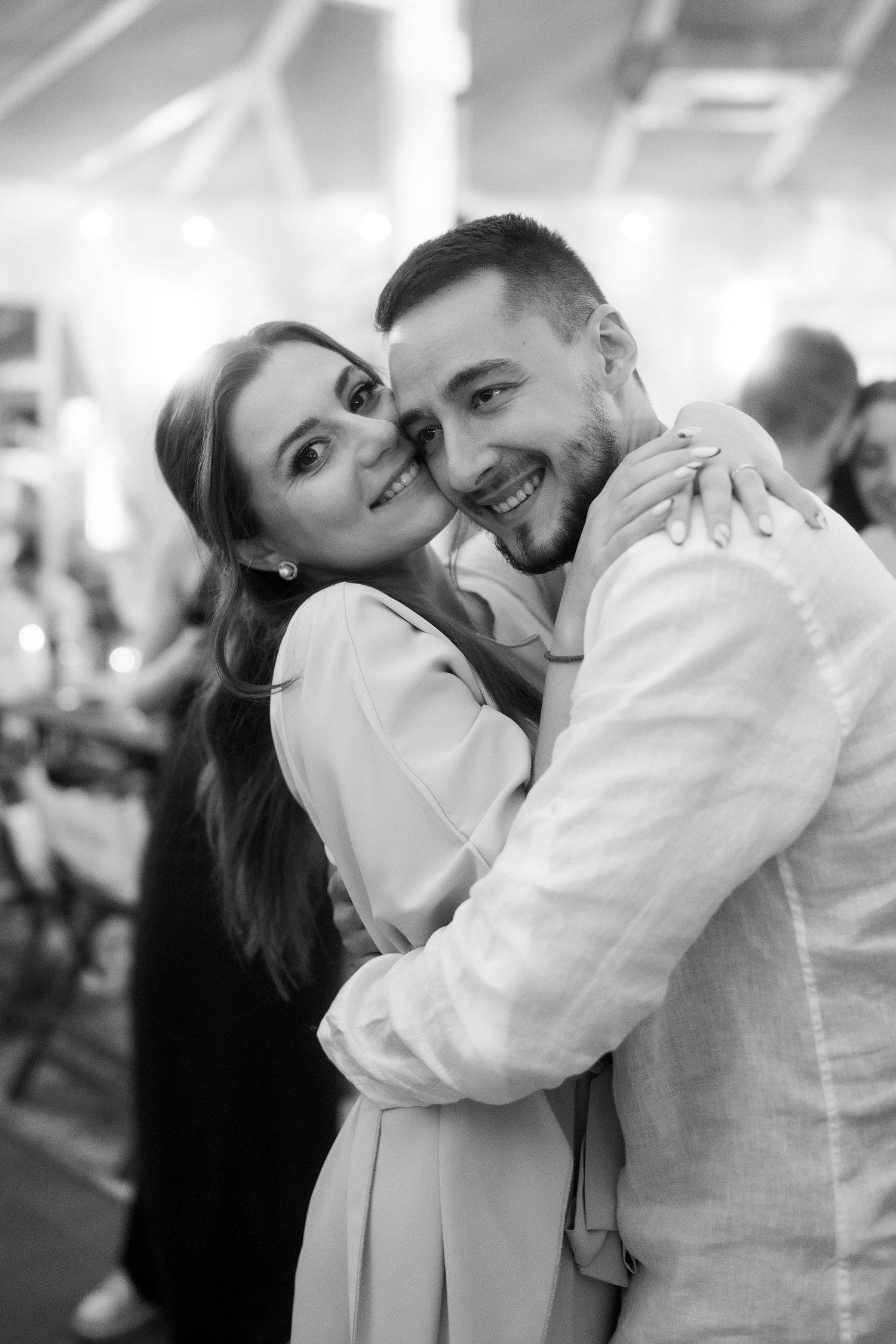 Black and white photo of a smiling couple embracing at an event, woman in white dress hugging man in light blazer.