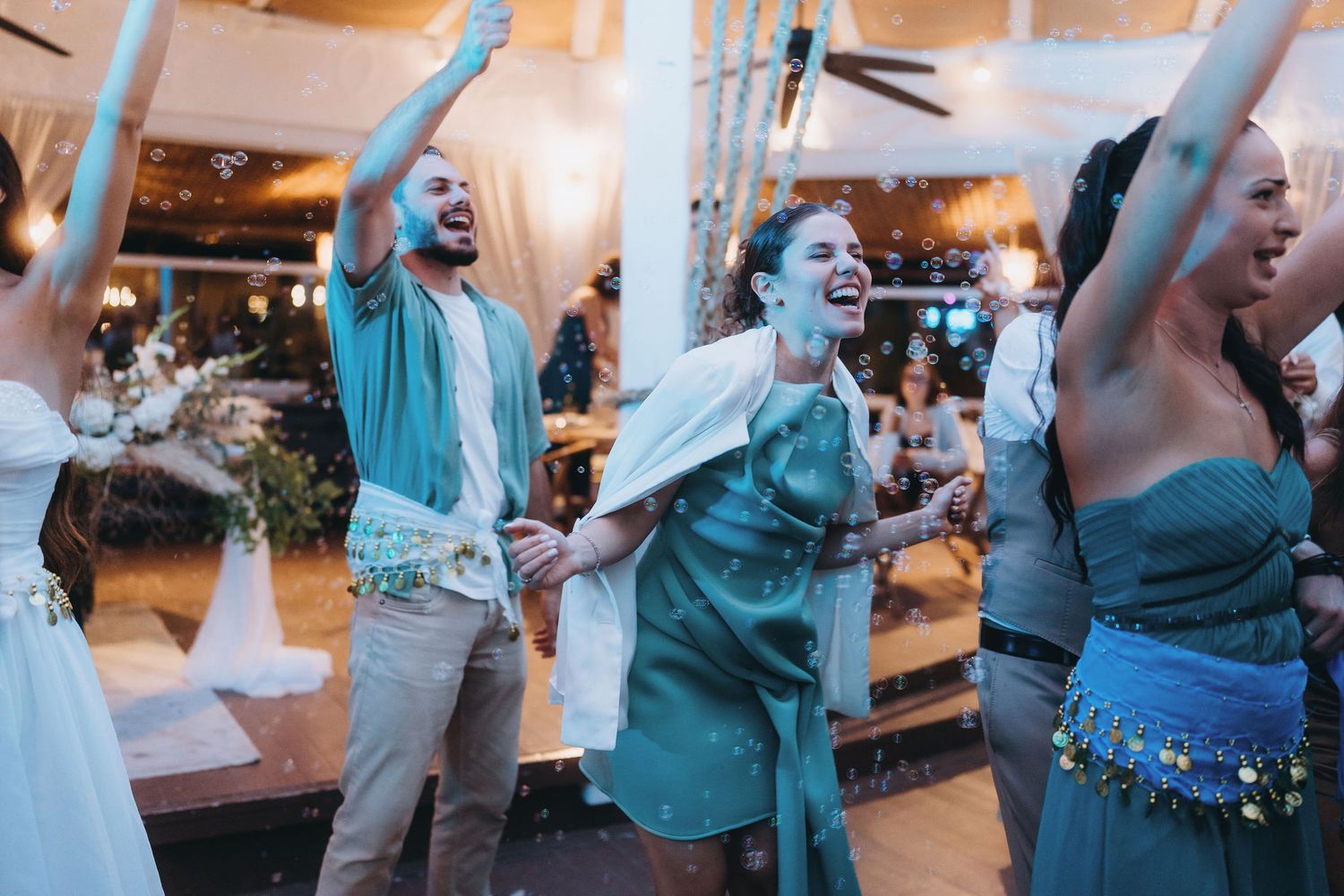 People dancing joyfully at a festive indoor celebration, wearing elegant teal and white outfits with confetti falling.