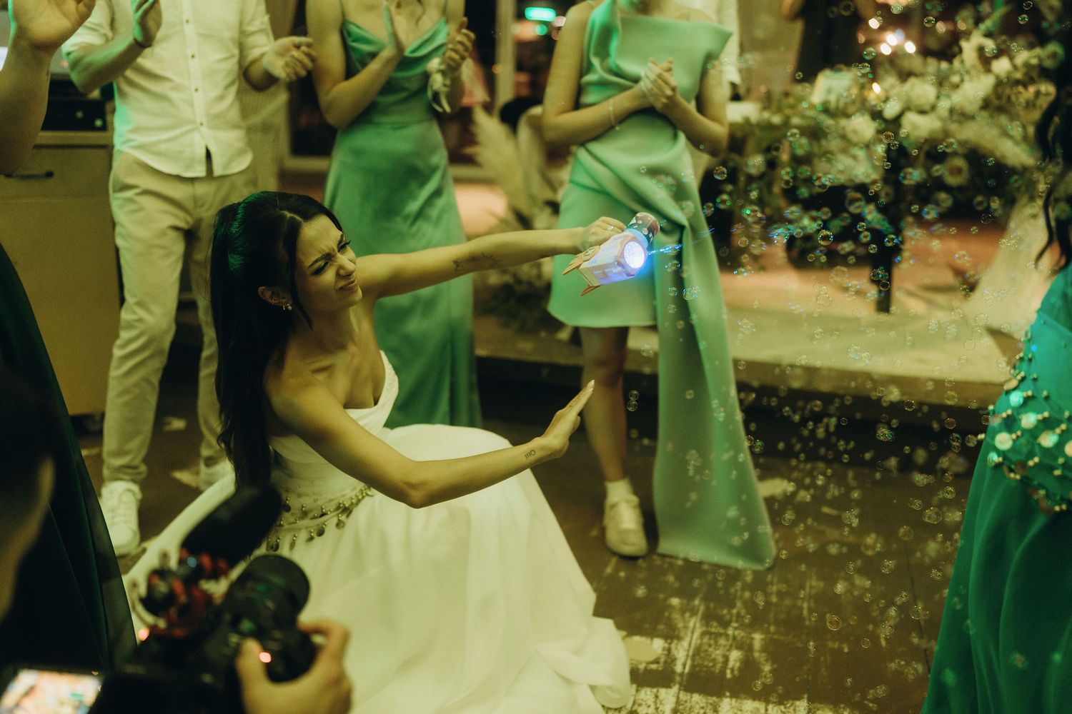 Bride in white dress kneeling on ground at outdoor wedding reception with guests in green dresses nearby.