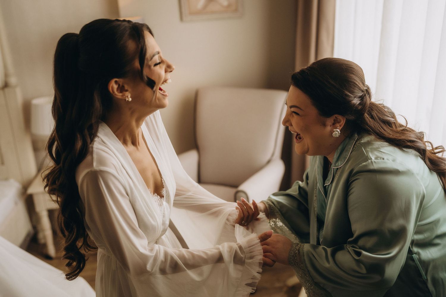 Two women in robes laughing joyfully together during bridal preparations in a bright, elegant room.