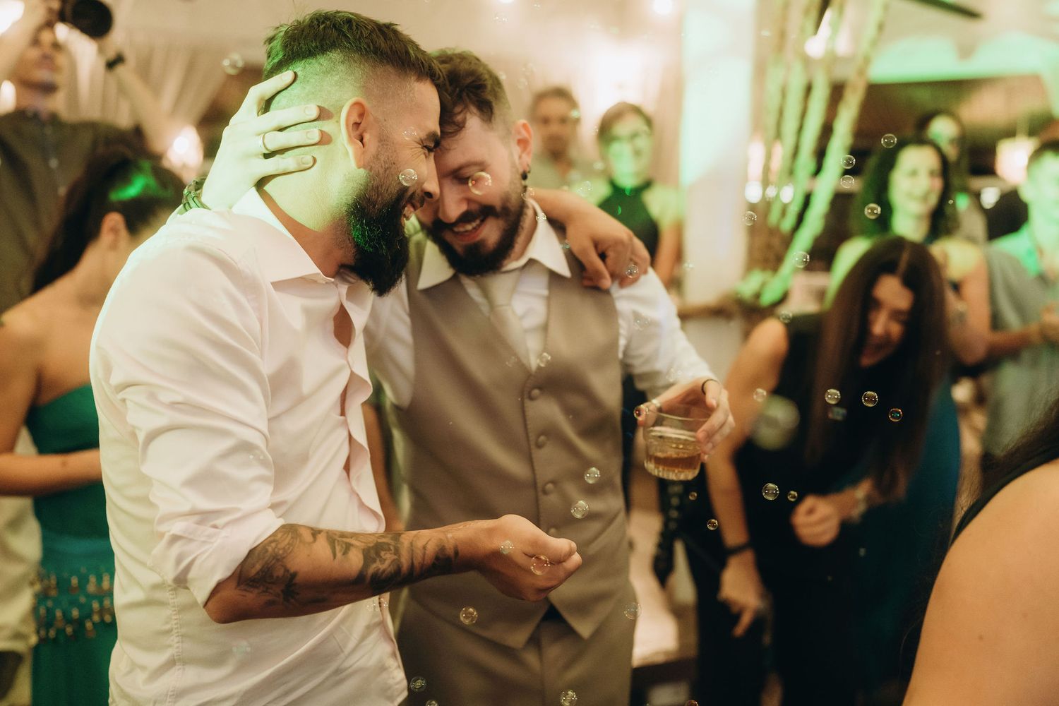 Two bearded men share a joyful embrace on a dance floor at a festive evening celebration.