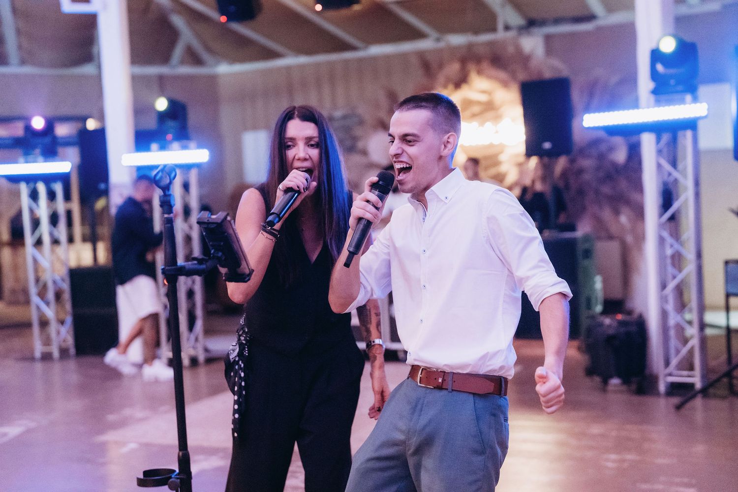 A man and woman singing together on stage with microphones at an indoor event venue.