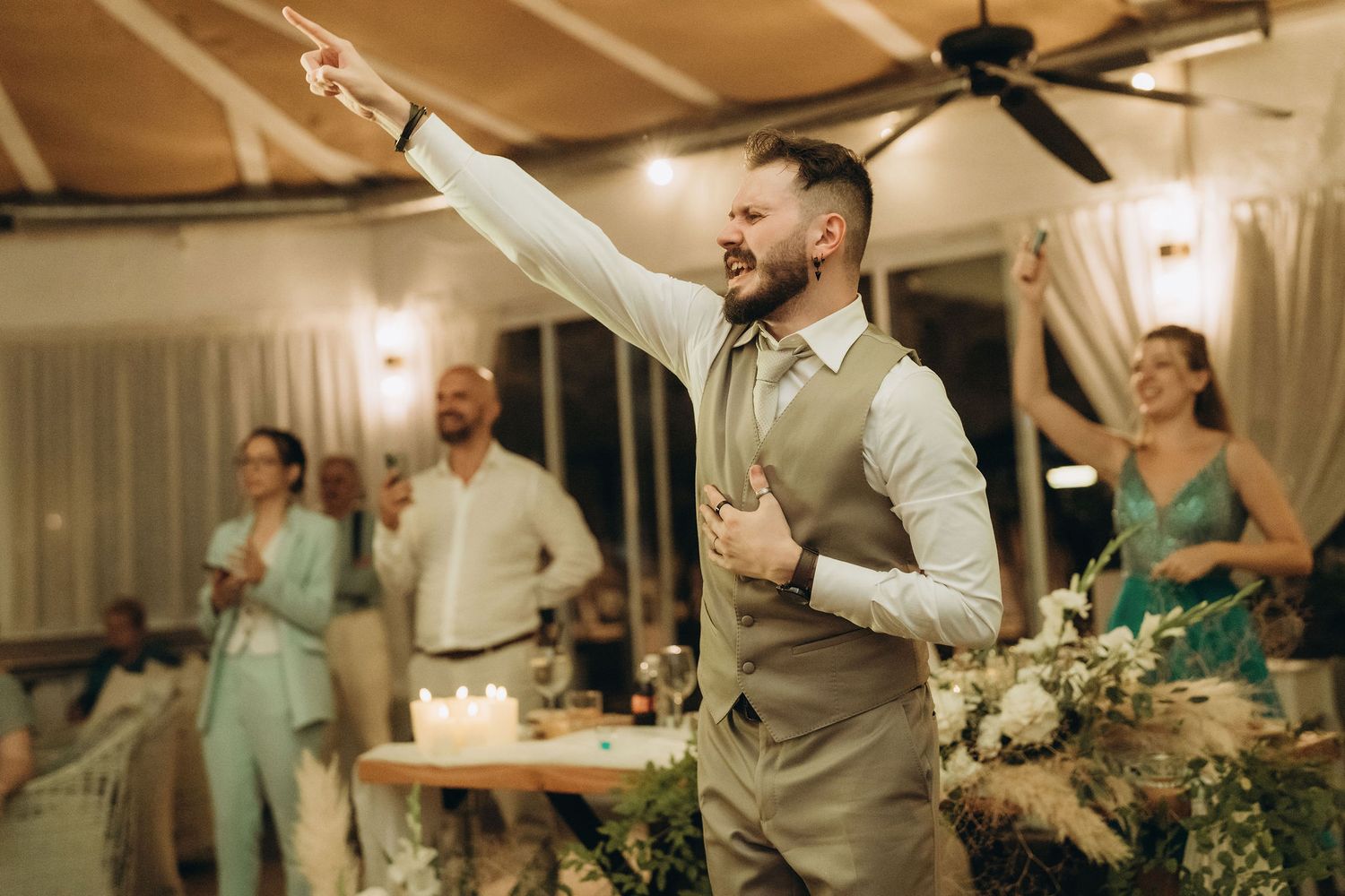 A groom in a gray vest celebrates joyfully at a wedding reception, pointing and laughing with guests in the background.