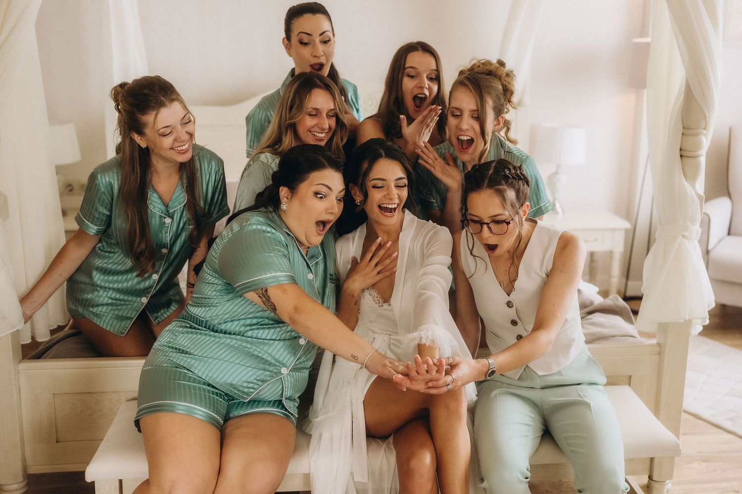 Excited bridesmaids in green satin pajamas admiring engagement ring with bride in white robe on bed.
