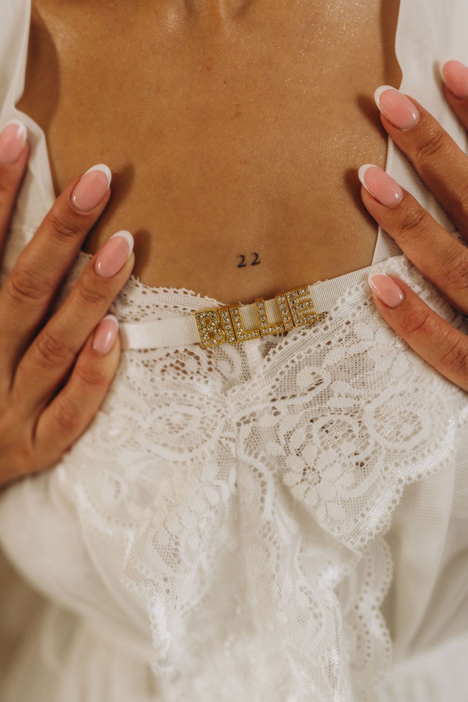 Close-up of a woman's midsection with a '22' tattoo, French manicure nails, and white lace lingerie with gold belt.
