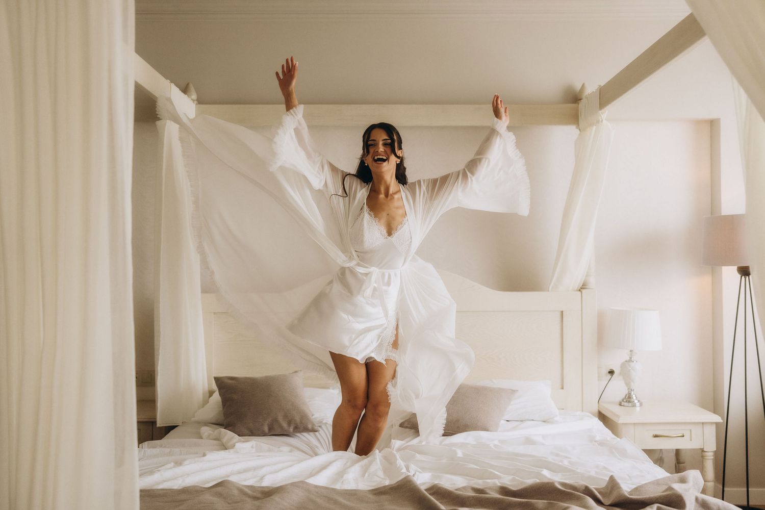 Joyful bride in white robe jumping on canopy bed with arms raised in bright airy bedroom.