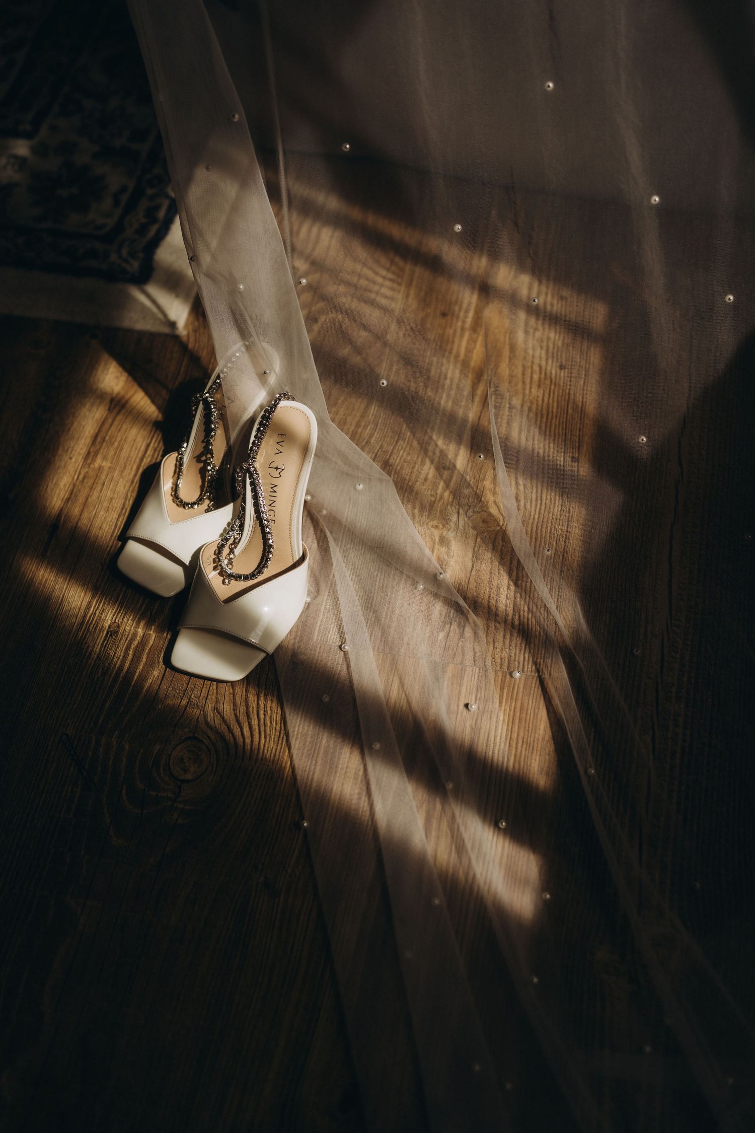 Elegant white bridal heels with chain detail resting on hardwood floor beside a flowing wedding veil in soft sunlight.